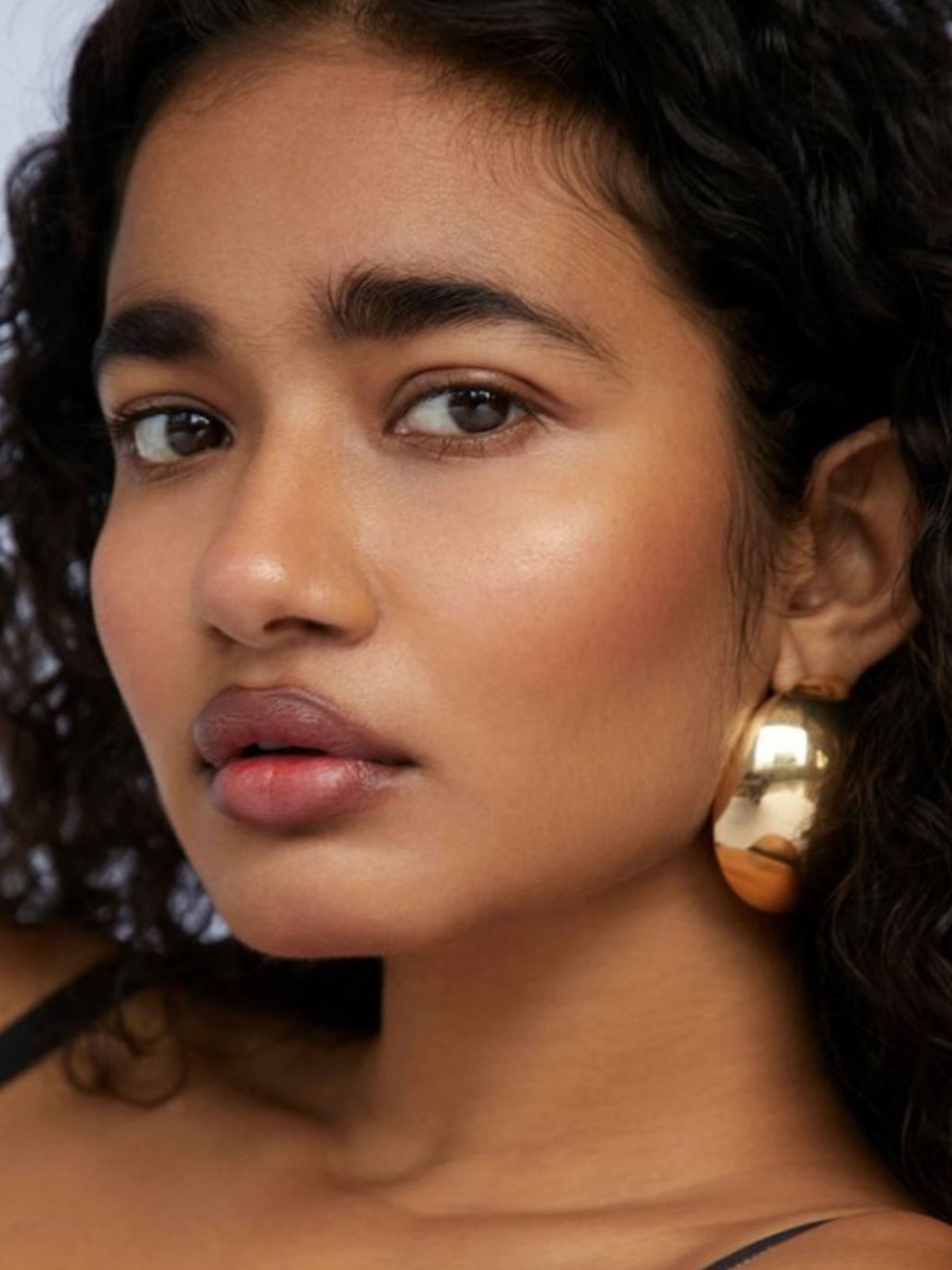 Woman with dark curly hair, a glowing bronzed cheek, nude lips and a large gold earring