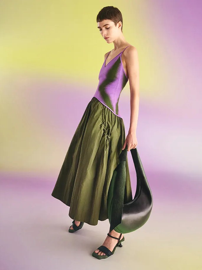Model in a purple knit top and olive skirt with a sculptural bag in a gradient studio