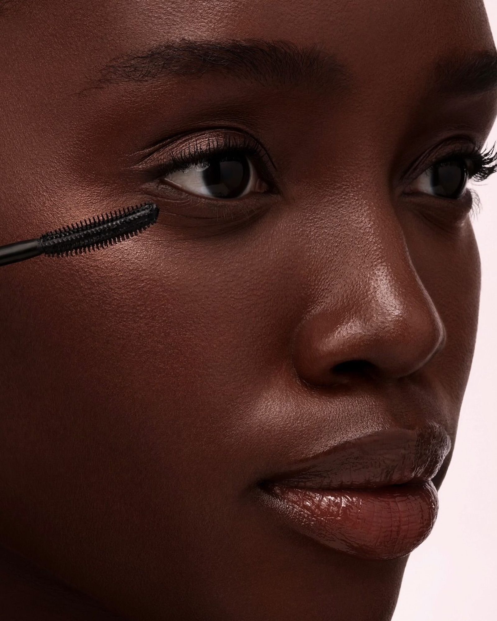 Close-up of a woman's face in soft light with a mascara wand near her lower lashes and glossy brown lips