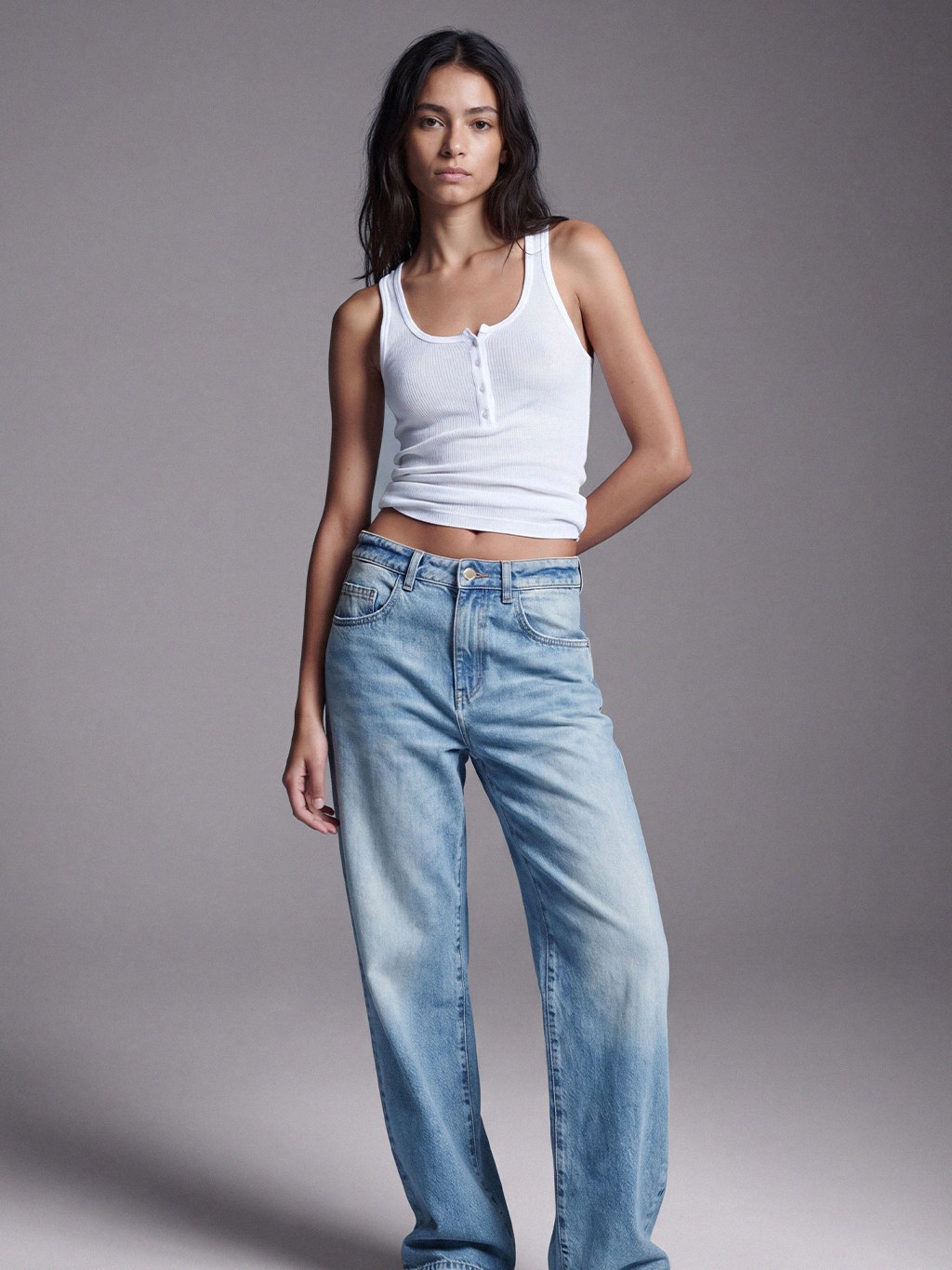 Model in wide light-wash jeans and a white tank against a gray studio backdrop.