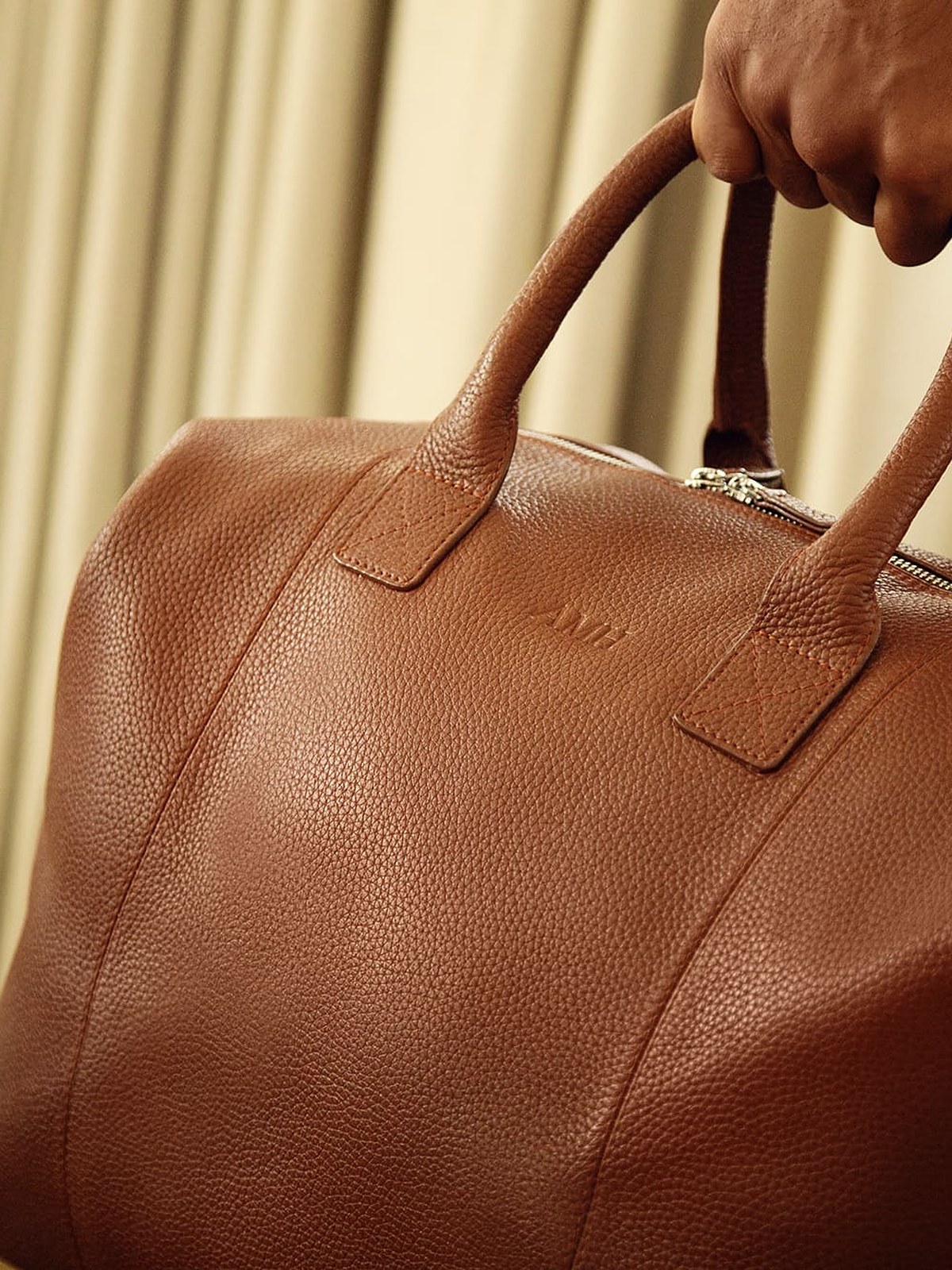 Hand carrying a cognac pebbled leather weekender bag against cream curtains