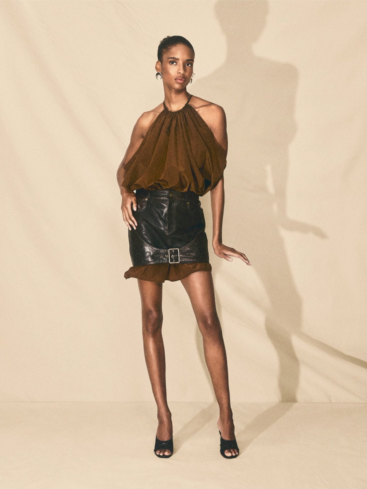 Model in a brown halter top and black leather mini skirt on a peach backdrop