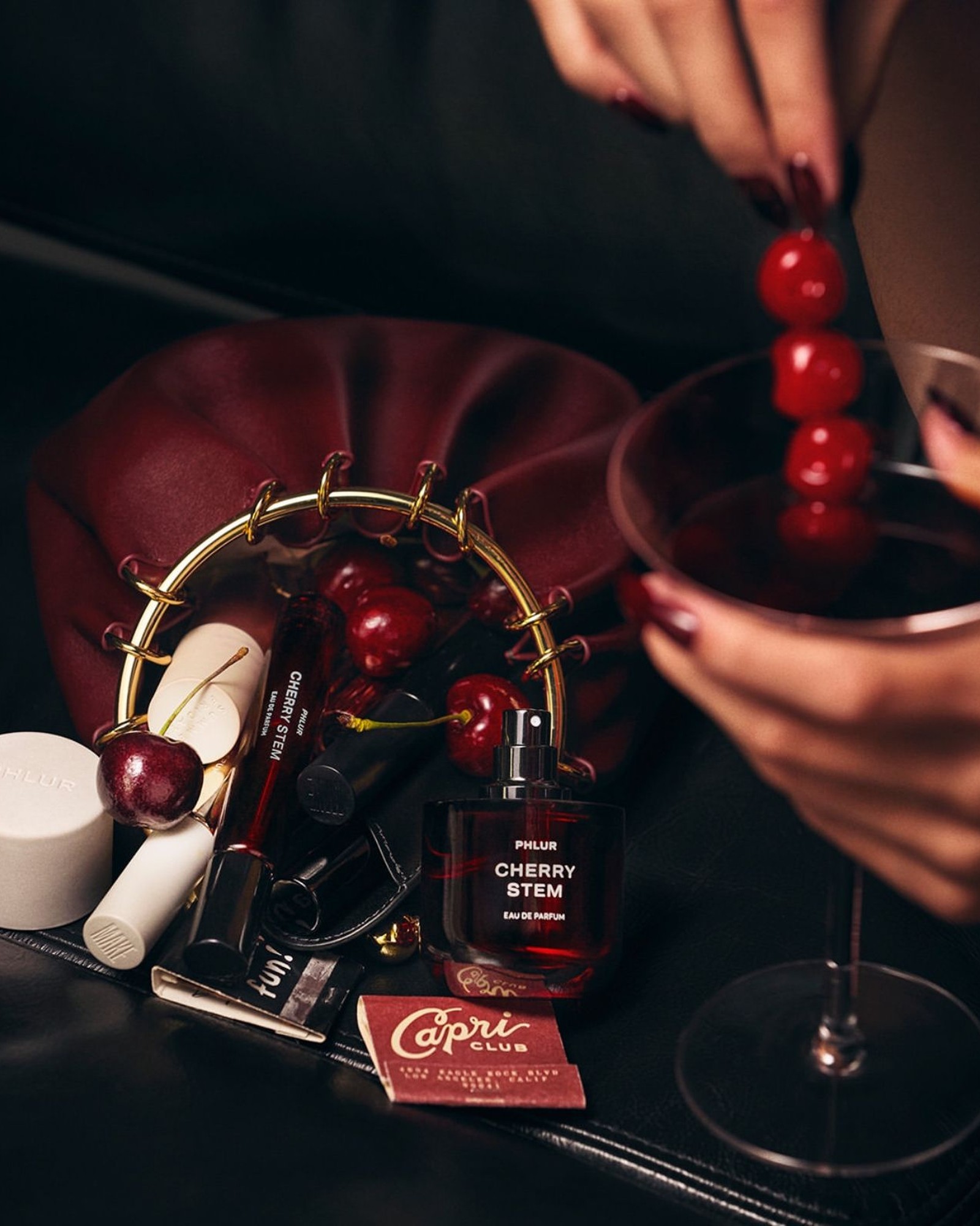 Bottle of PHLUR Cherry Stem eau de parfum among fresh cherries, lip products and a matchbook beside a hand holding a cocktail with cherries