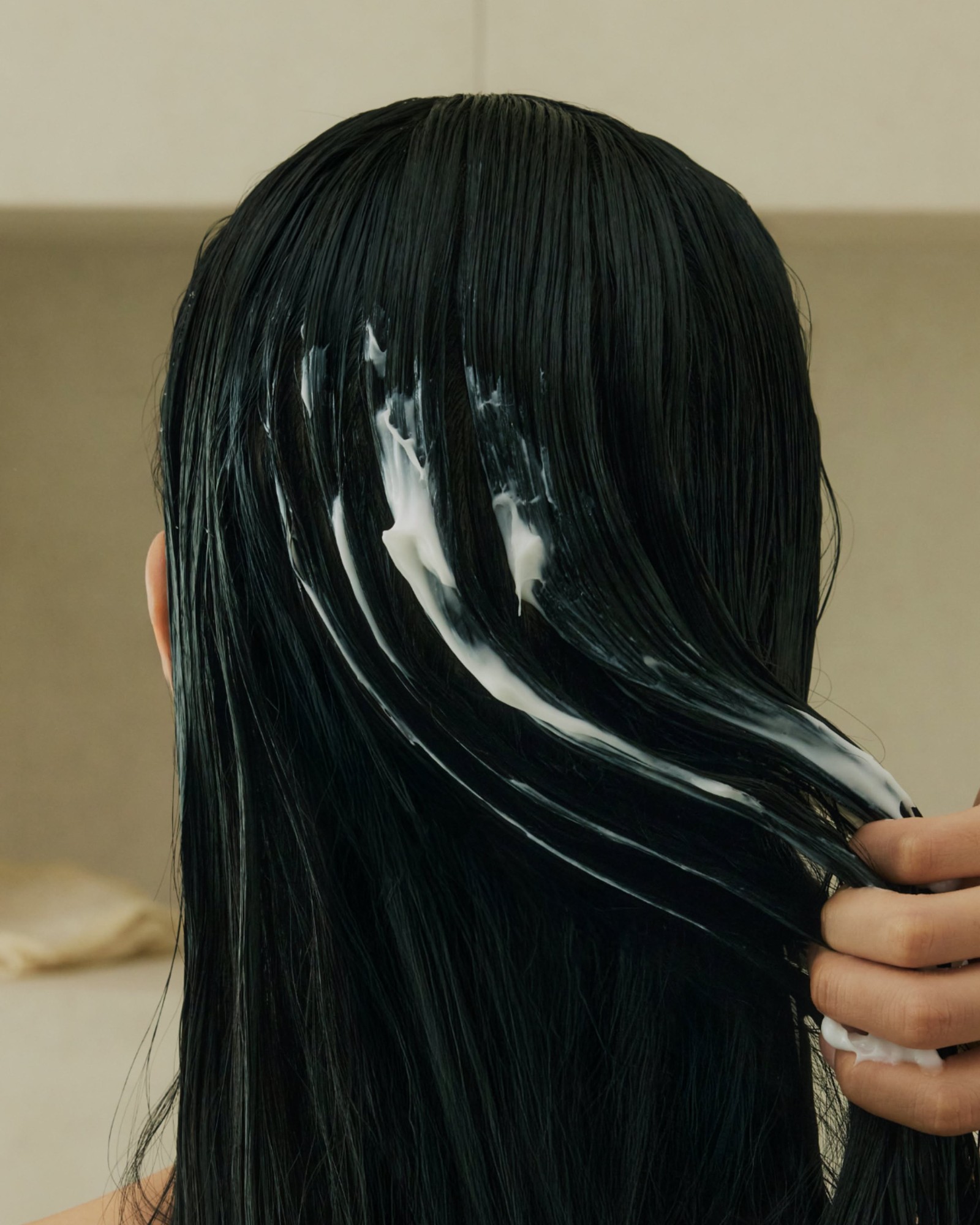 Back of a woman's long wet black hair with white conditioner smoothed through it by one hand