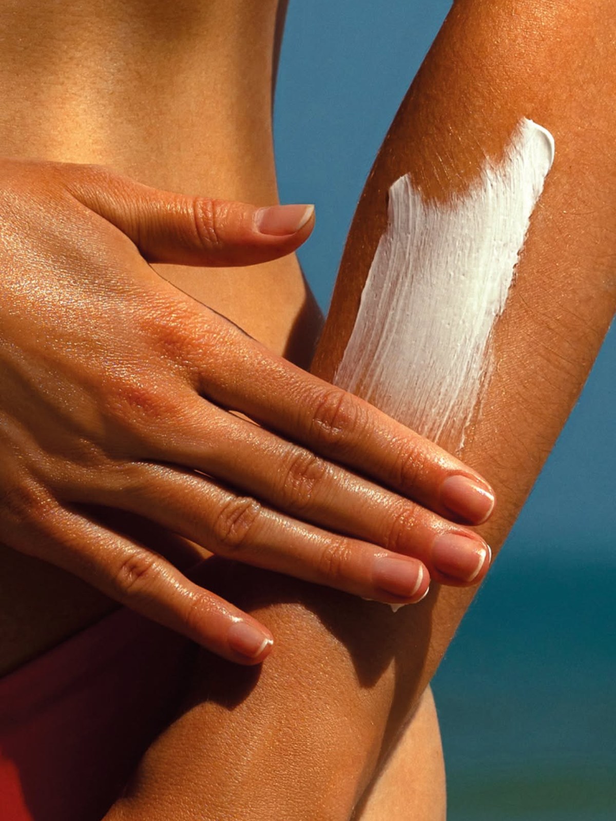 Close-up of a tanned hand spreading a stripe of white sunscreen down a forearm against a blue sea