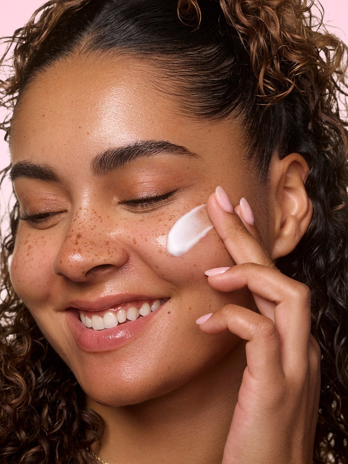 Smiling curly haired model smoothing cream onto a freckled cheek