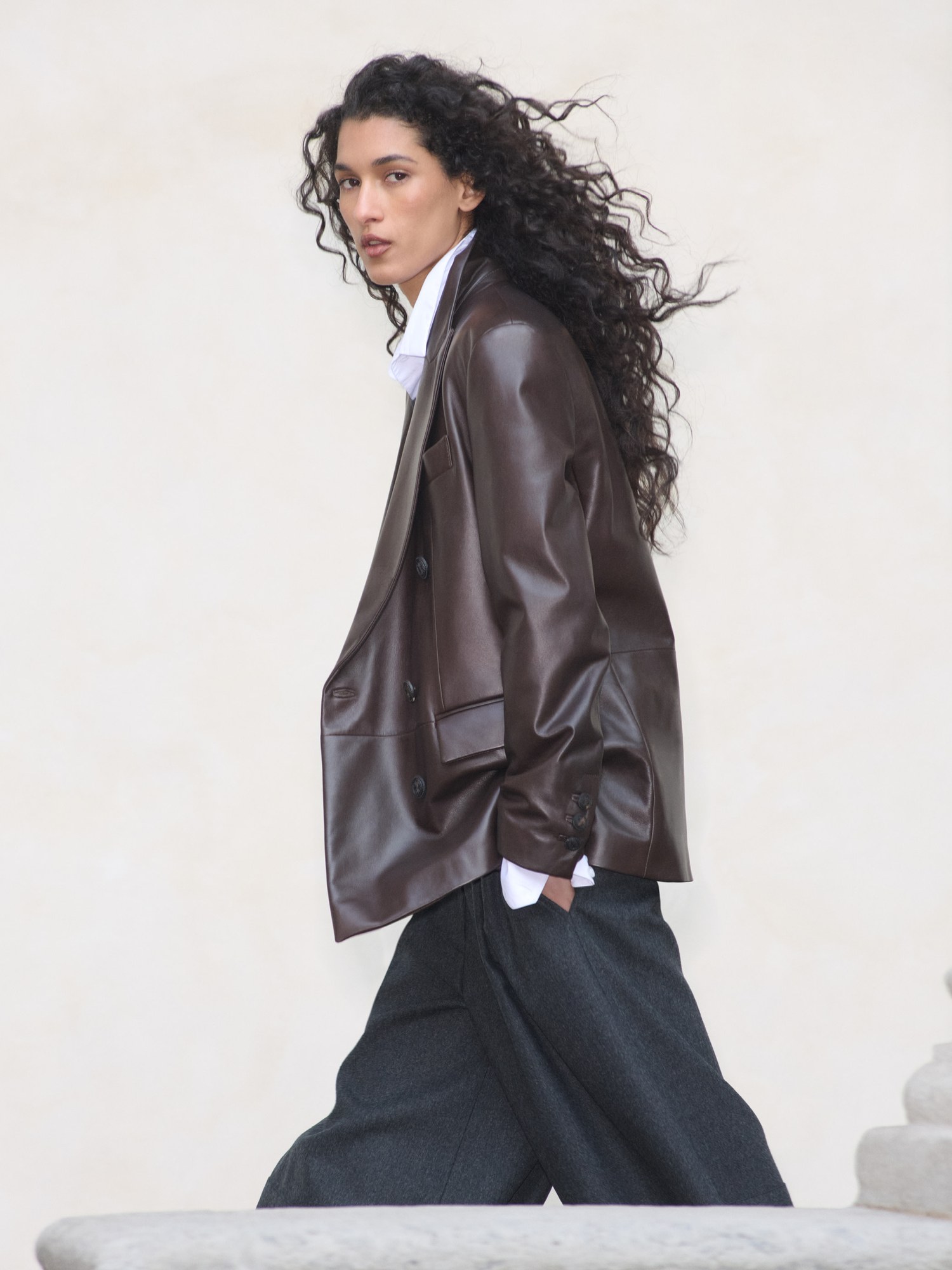 Model with windswept curly hair in an oversized brown leather jacket and wide grey trousers