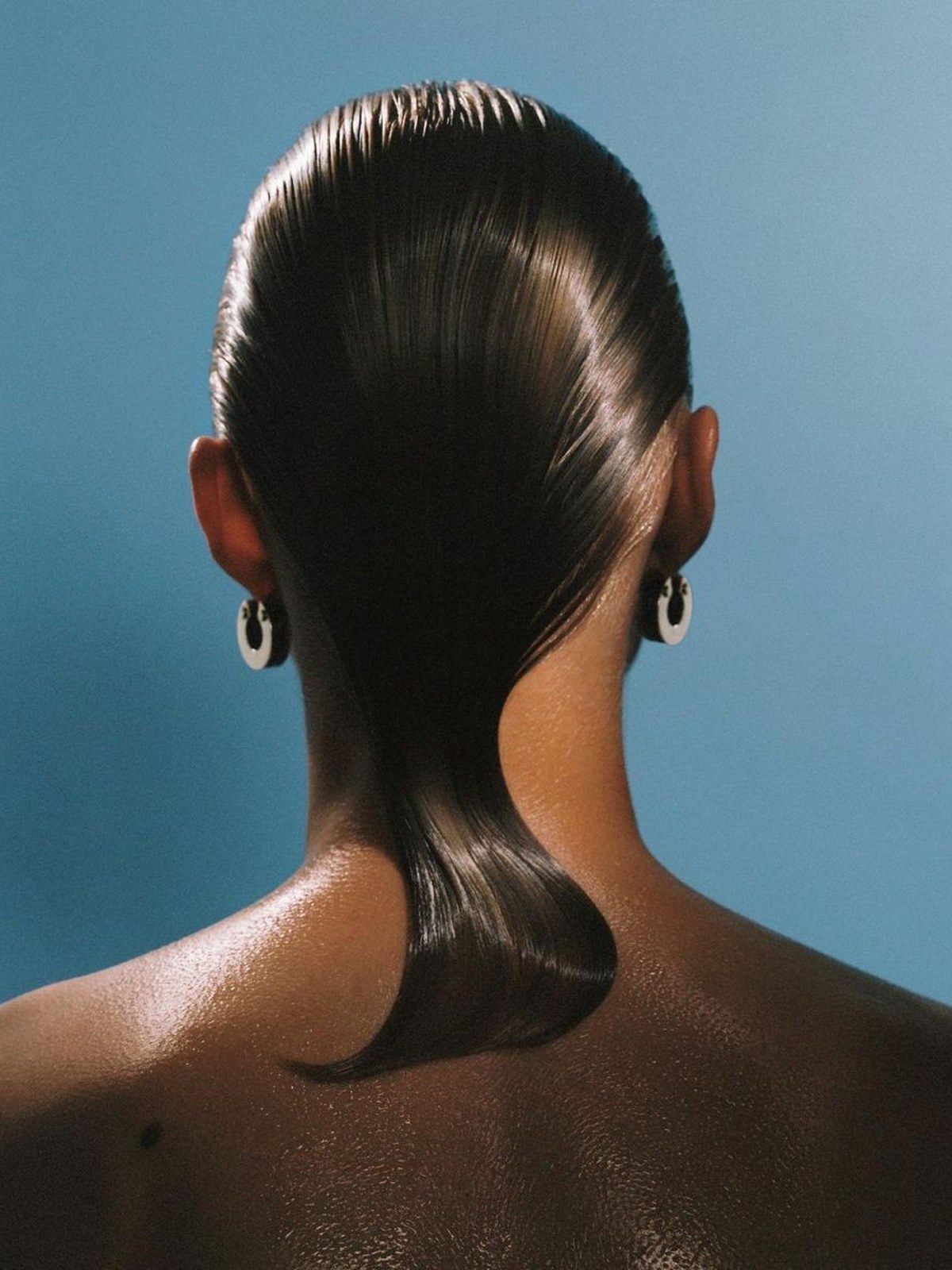 Back of a head with a glossy dark slicked-back low bun and a gold ridged earring, slightly blurred