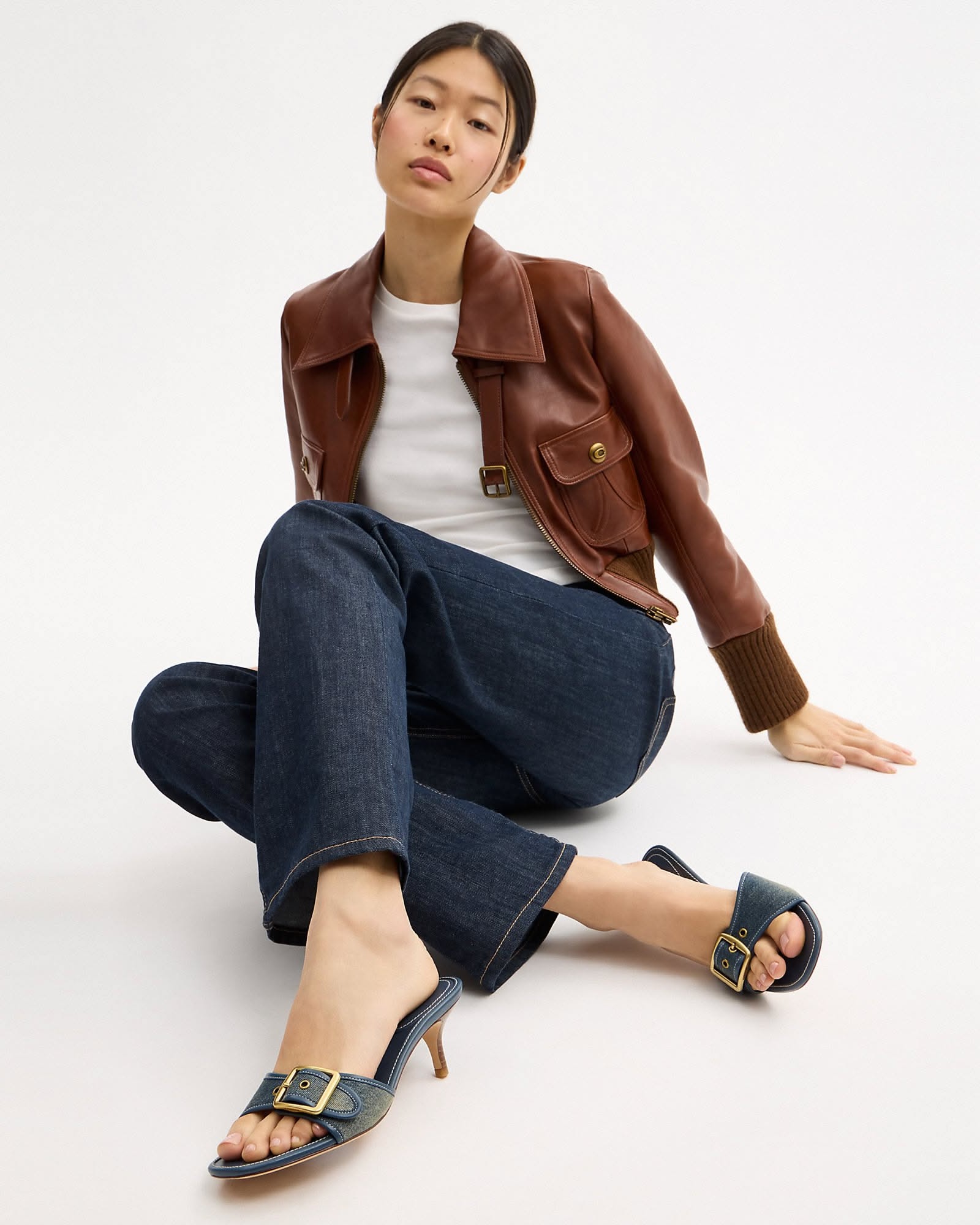 Woman sitting on a white floor in a brown leather jacket, white tee and dark jeans with black Coach Margot buckle sandals