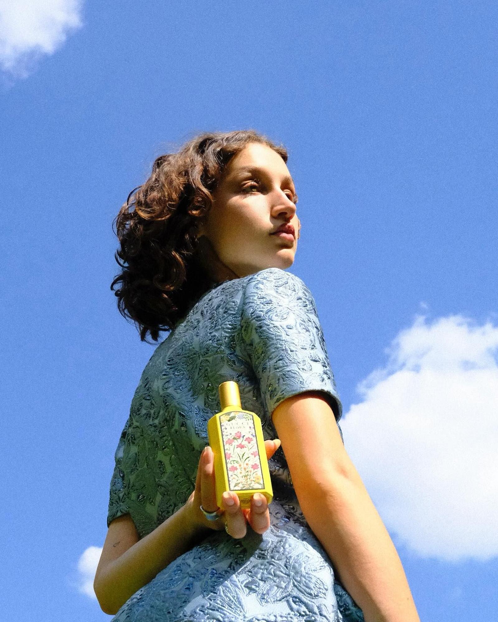 Woman with curly brown hair in a blue jacquard top holding a yellow Gucci Flora bottle against a bright blue sky