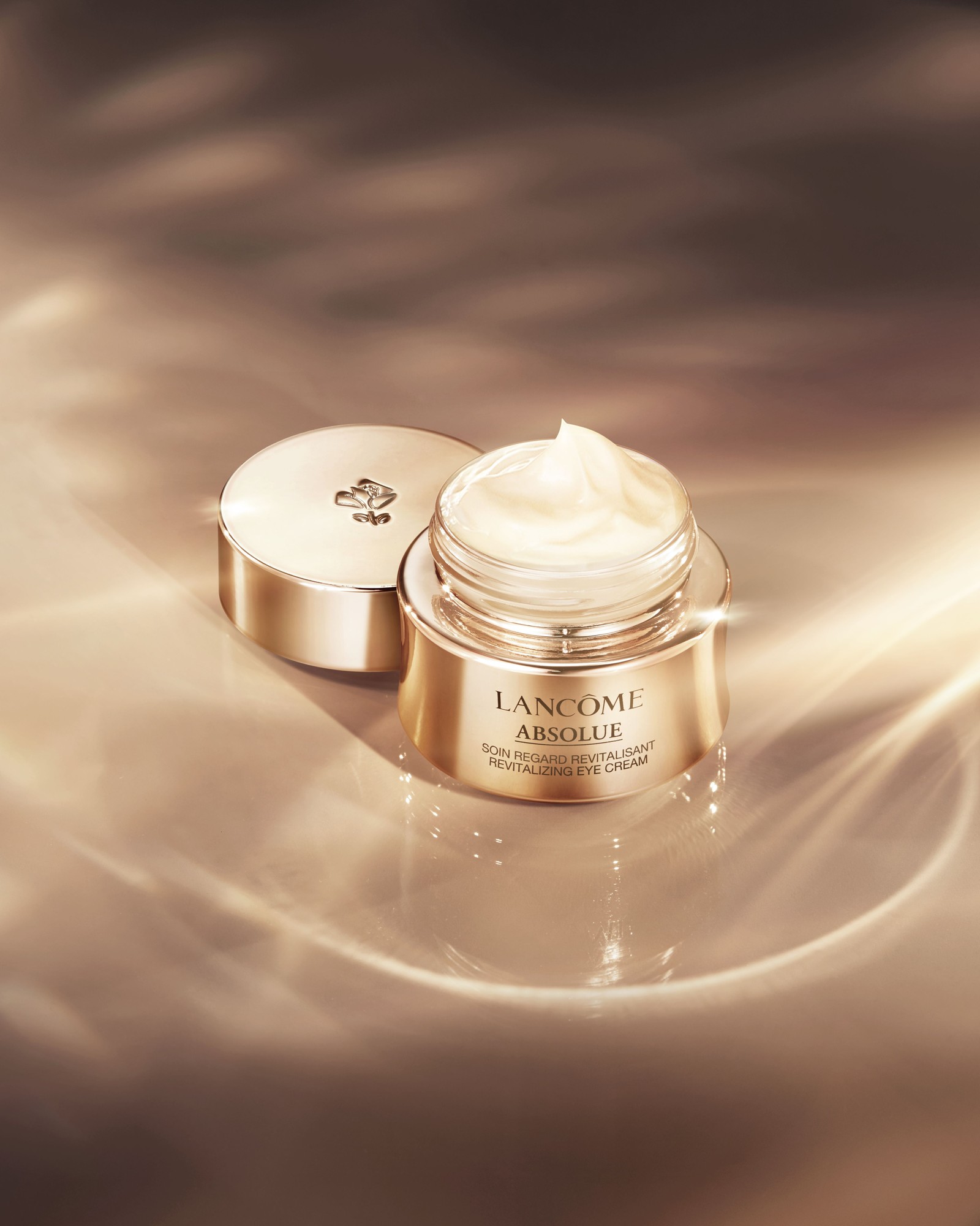 Open gold jar of Lancôme Absolue Revitalizing Eye Cream with its lid beside it, on a golden reflective surface