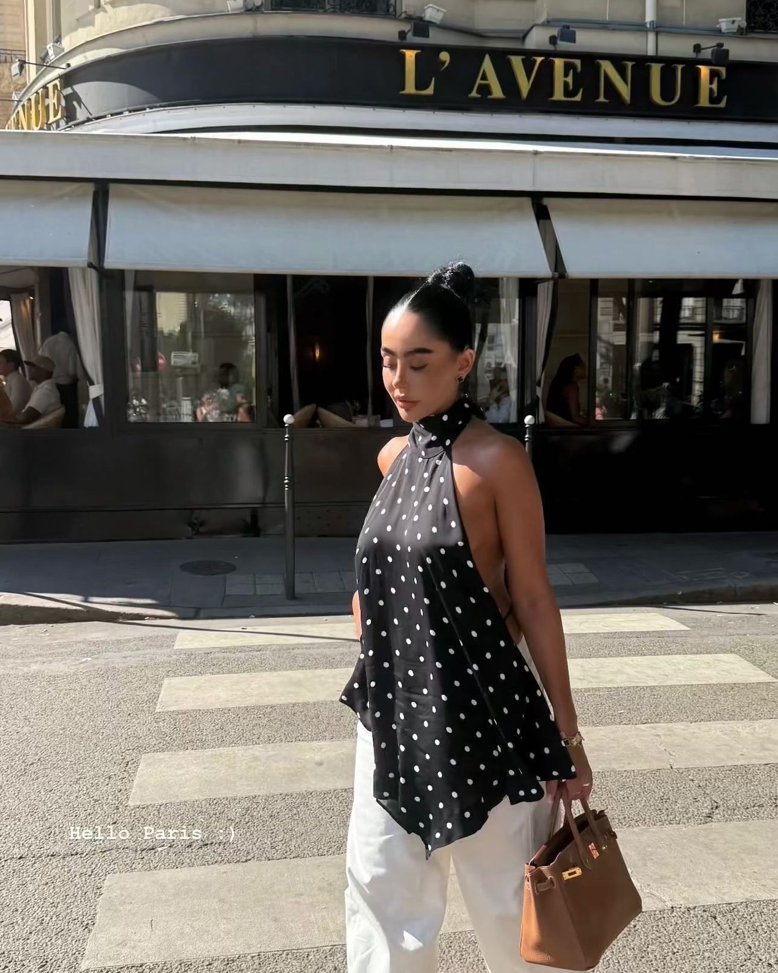 Woman crossing a street in a black and white polka dot halter top, white wide-leg trousers and a tan top-handle bag