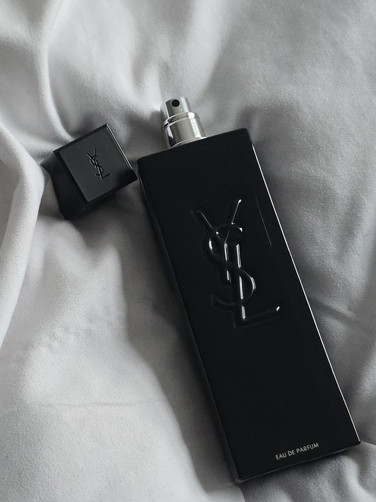 Black YSL cologne bottle with its cap removed lying on a white sheet