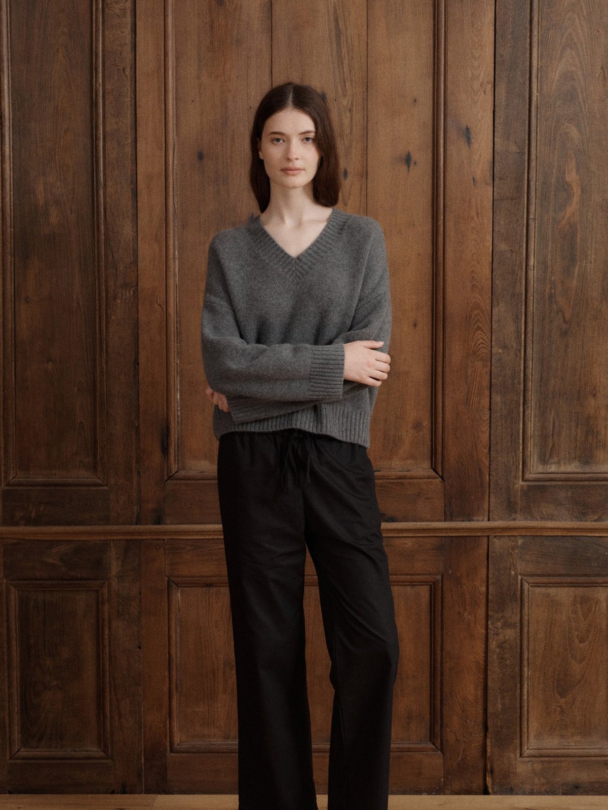 Model in a grey V-neck sweater and black trousers against wood paneling