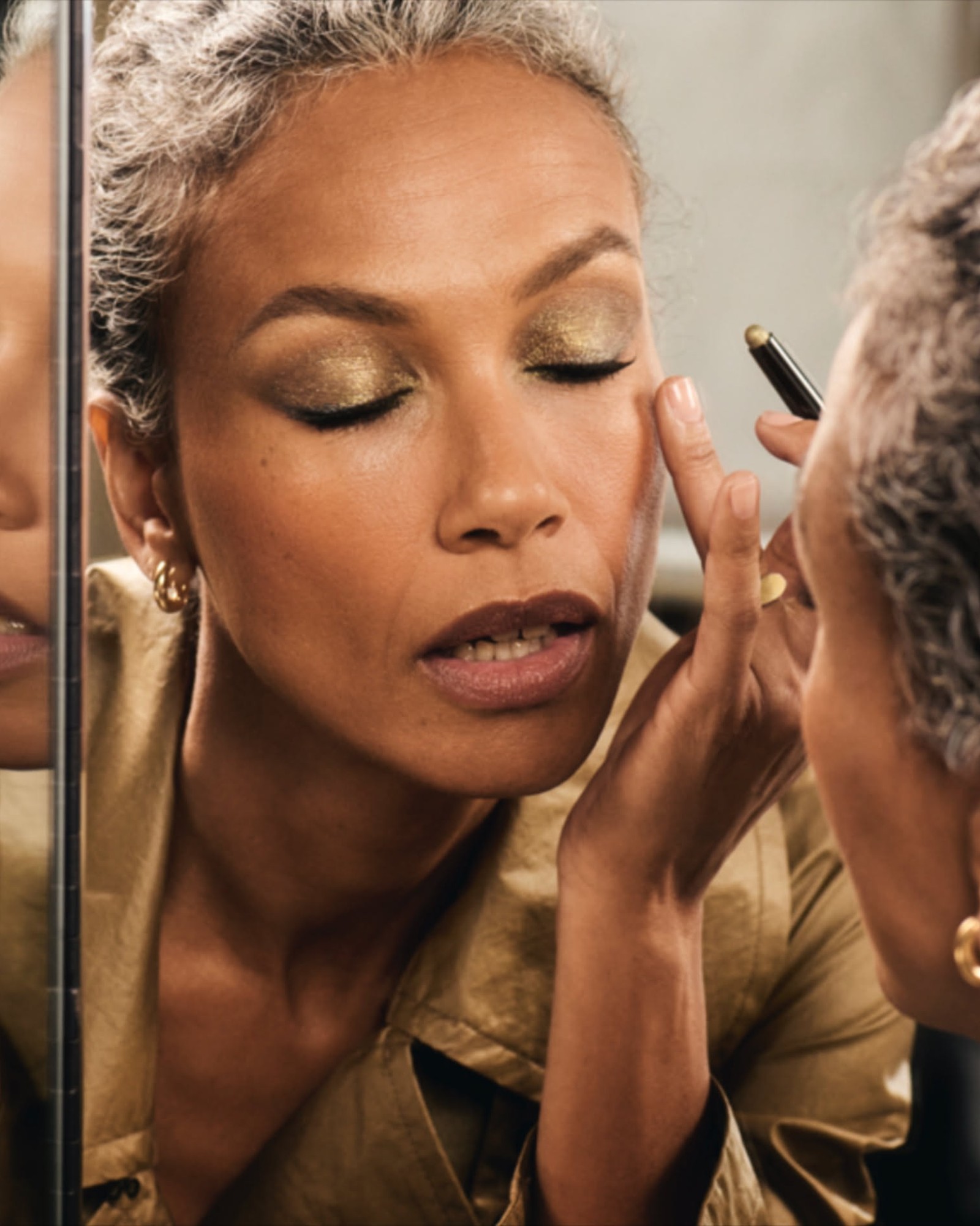 Woman with grey hair pulled back applying gold shimmer eyeshadow in a mirror, wearing a gold jacket