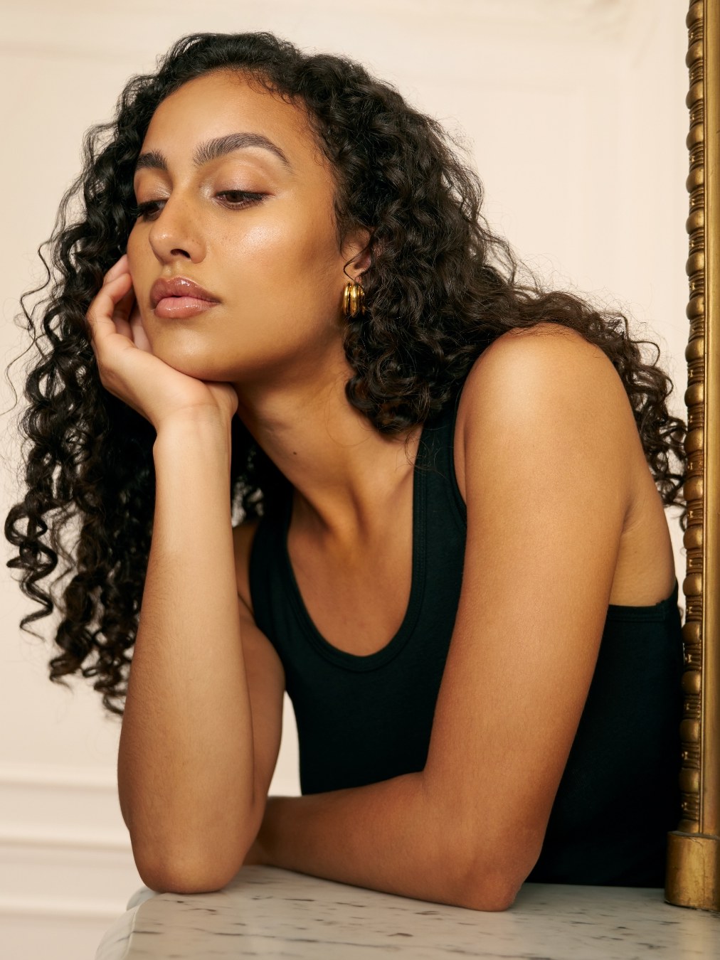 Woman with defined curls resting her chin on one hand in a cream-toned studio