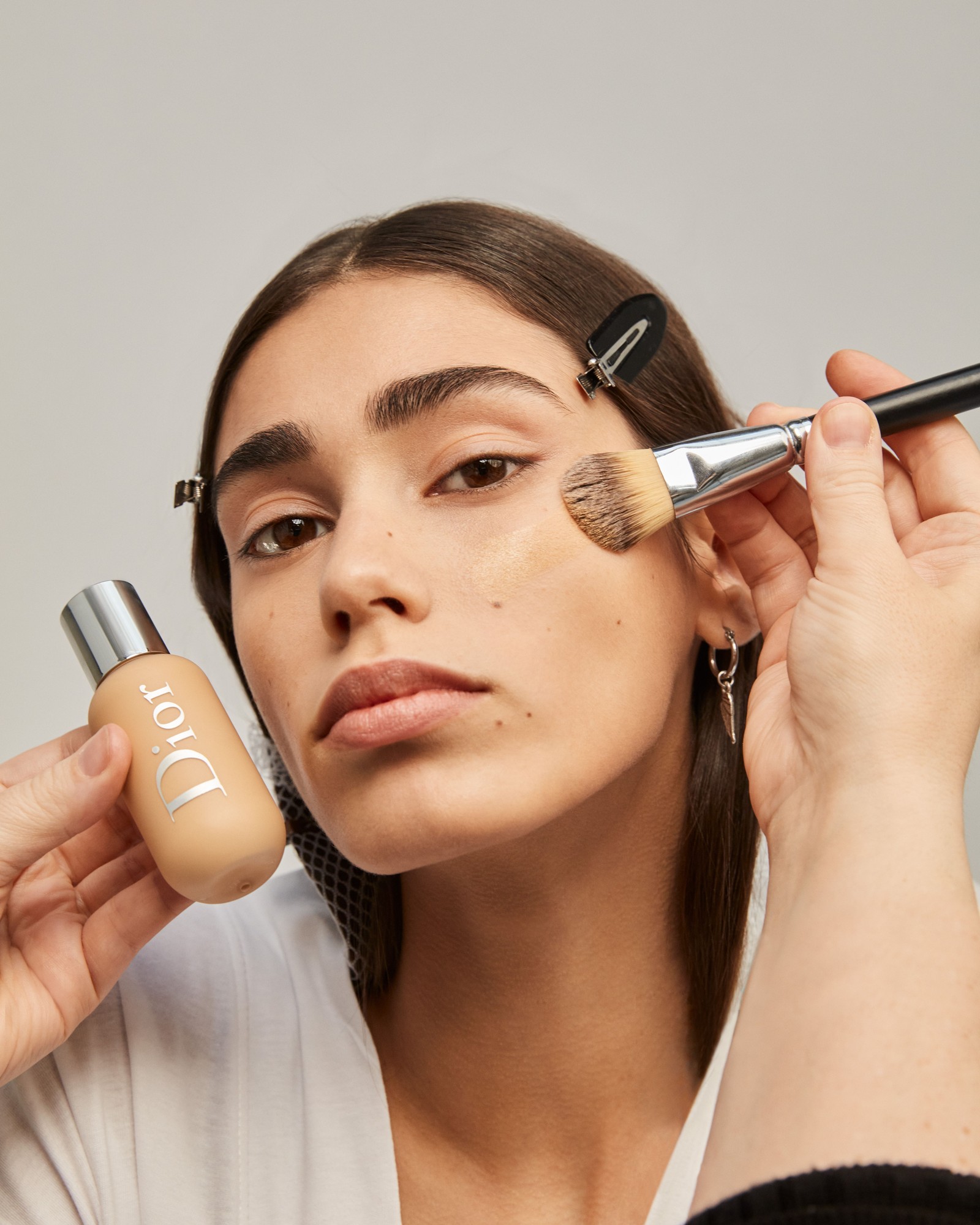 Model with brown eyes having foundation brushed onto her cheek from a Dior Backstage bottle, her hair clipped back in a white robe
