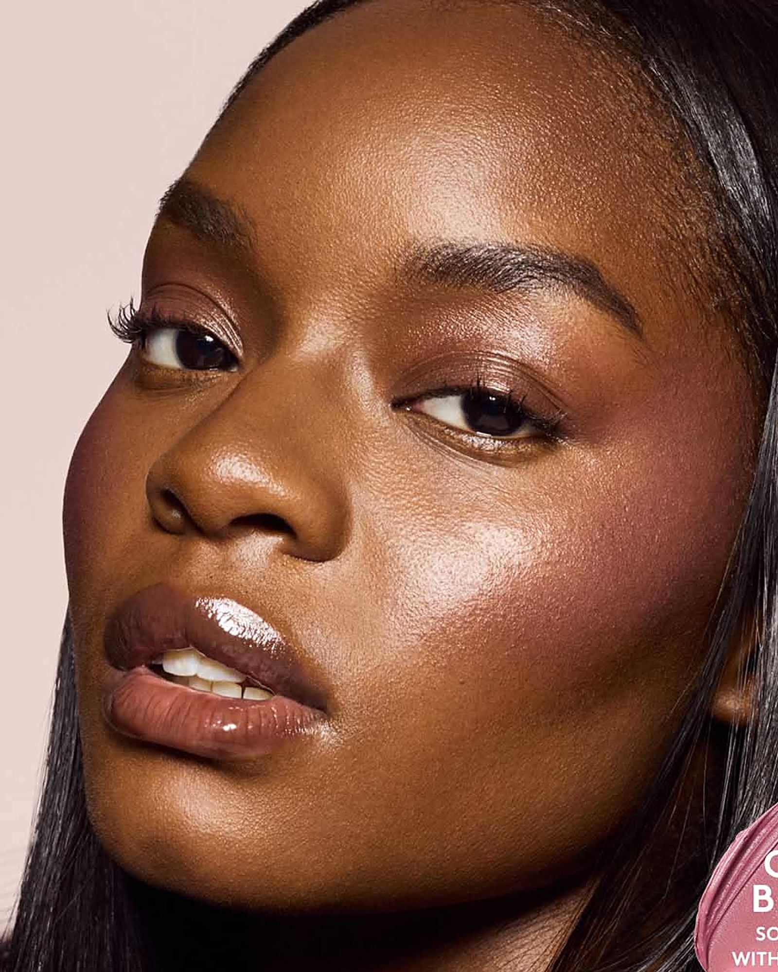 Close-up of a woman with deep skin, glowing cheeks and a glossy nude lip against a pale pink background