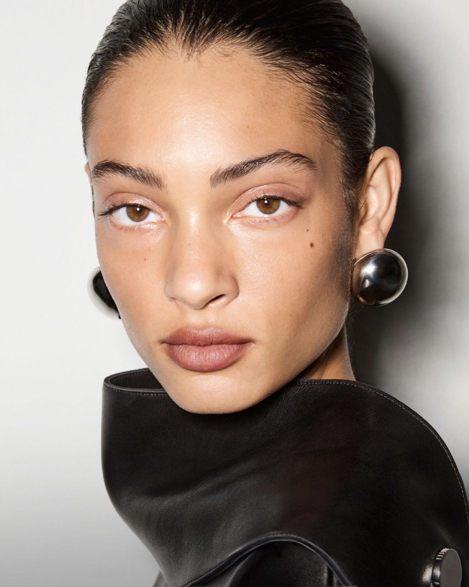 Close-up of a model with slicked-back hair, a natural glowing base, a soft nude lip and large silver ball earrings on a black leather collar