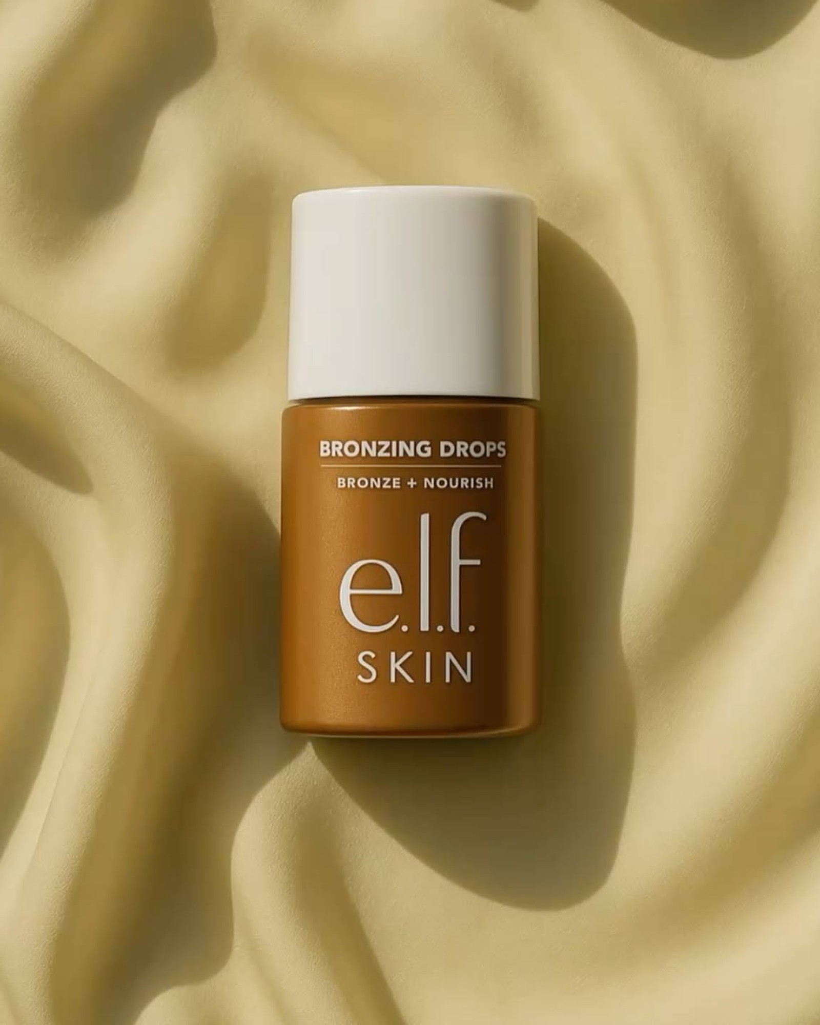 Bottle of e.l.f. SKIN Bronzing Drops with a white cap resting on soft pale yellow satin folds