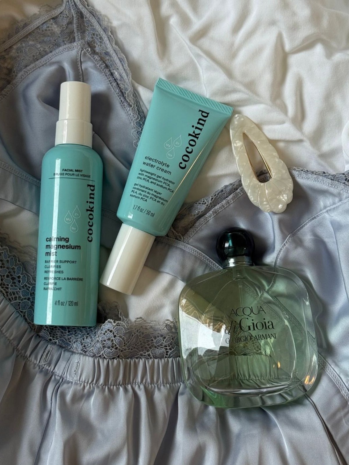 Cocokind Calming Magnesium Mist and Electrolyte Water Cream flat lay on blue satin with a pearl hair clip and an Armani Acqua di Gioia bottle