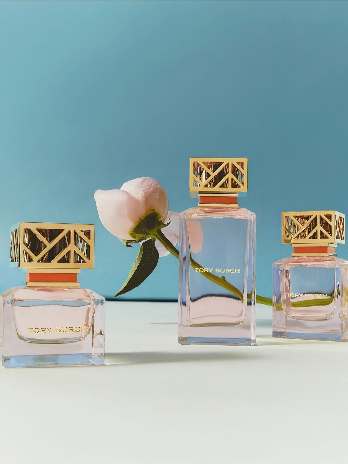 Three Tory Burch perfume bottles with gold geometric caps beside a pale pink peony against a blue backdrop