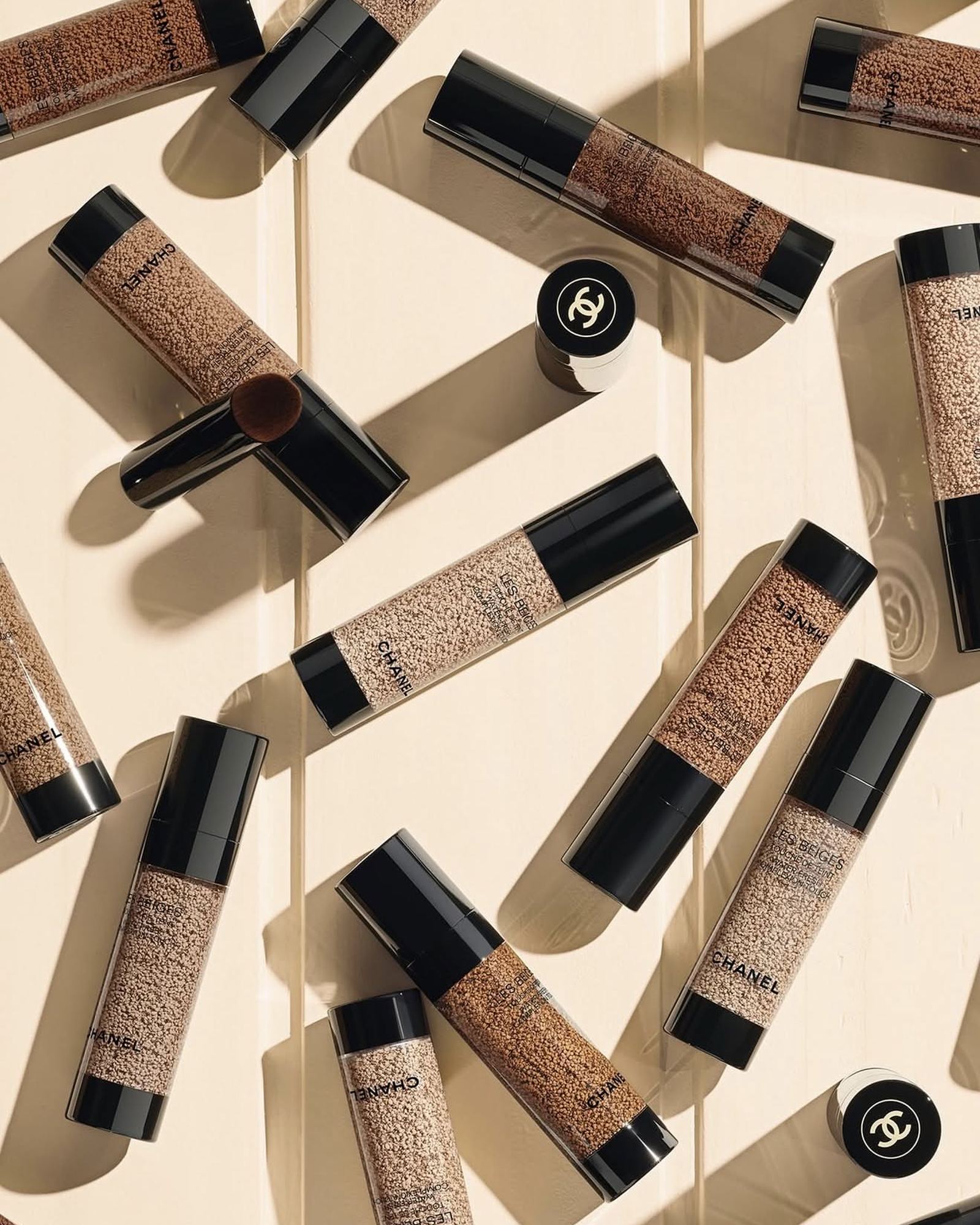 Overhead shot of many Chanel Les Beiges Water-Fresh Tint bottles in different shades scattered on a cream wooden surface, with small brushes and black Chanel caps among them.