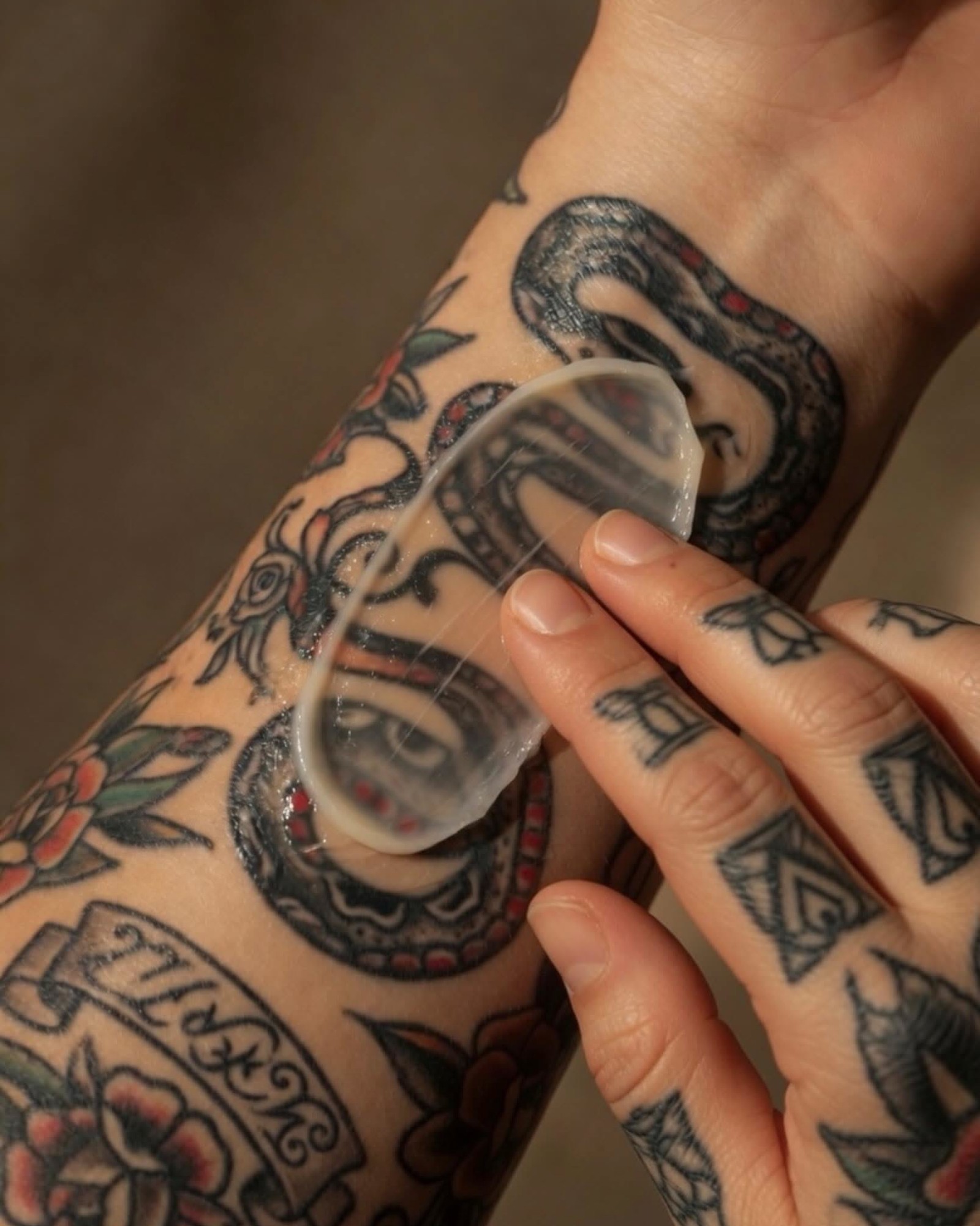 Forearm and fingers covered in traditional tattoos of a snake and roses, with a fingertip pressing a clear translucent gel sheet against the skin