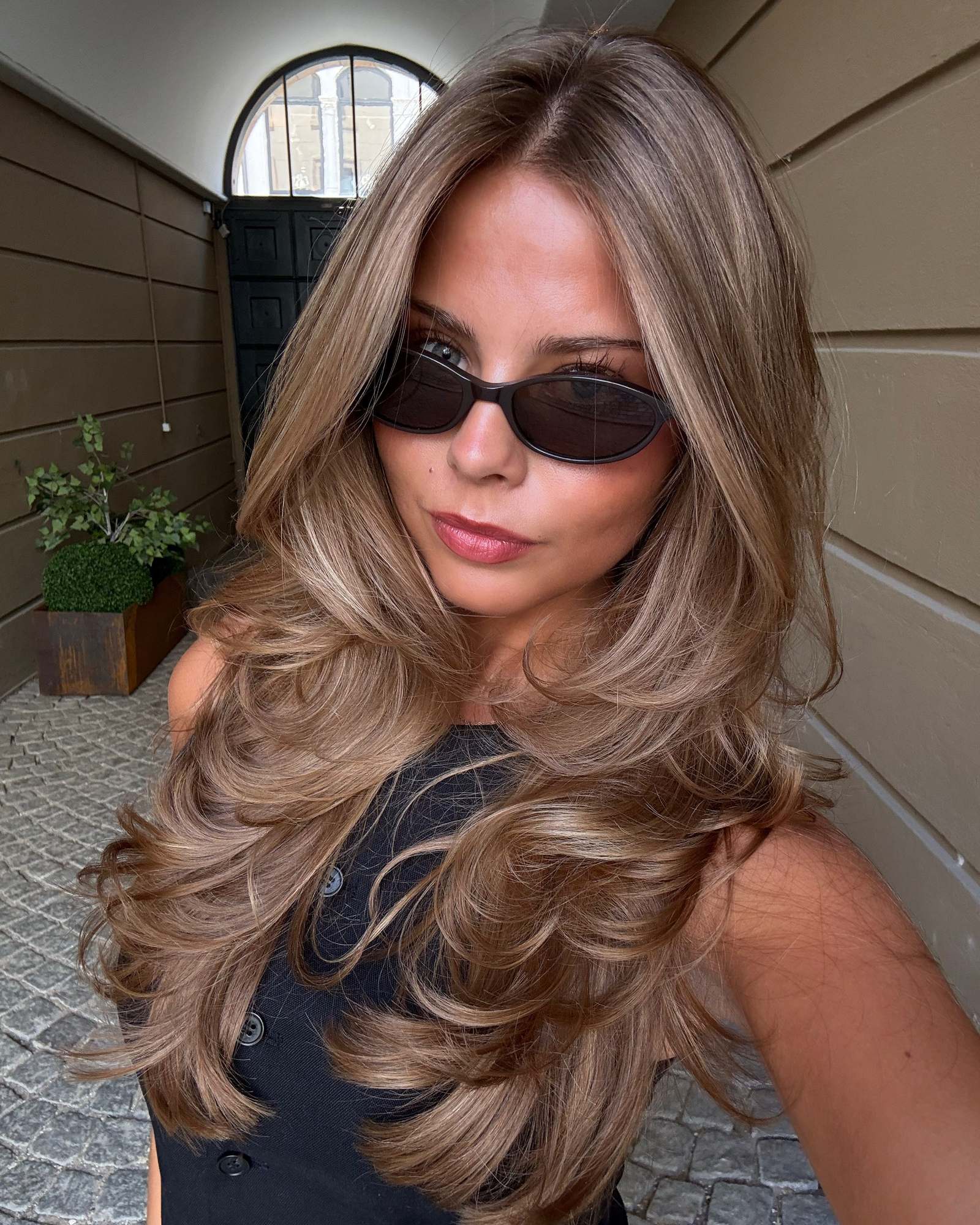 Woman in oval sunglasses and a black top taking a selfie with long layered light brown hair in loose waves