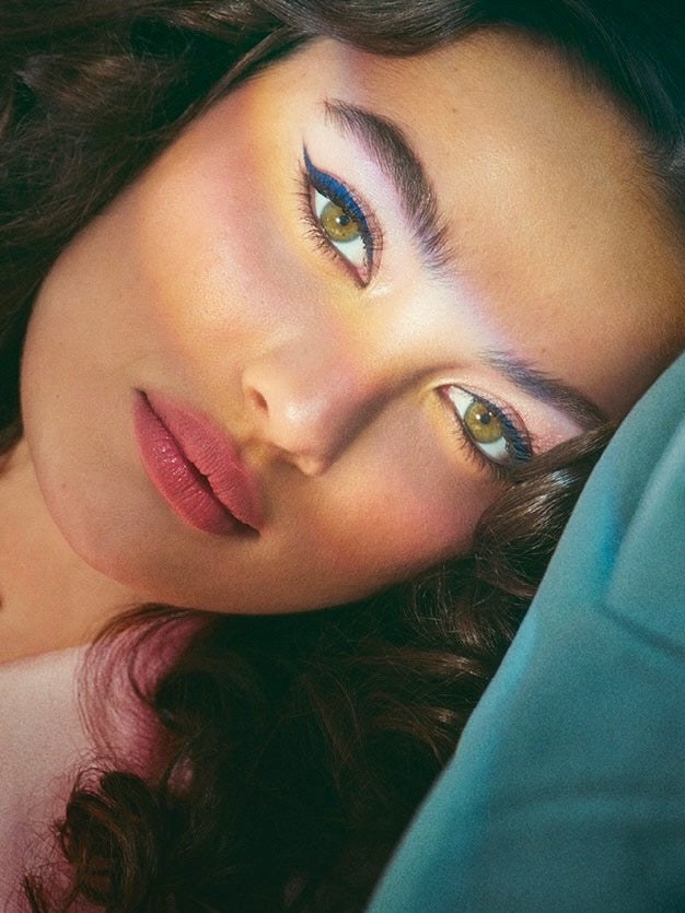 Model with luminous eye makeup and dark curls against a deep teal backdrop