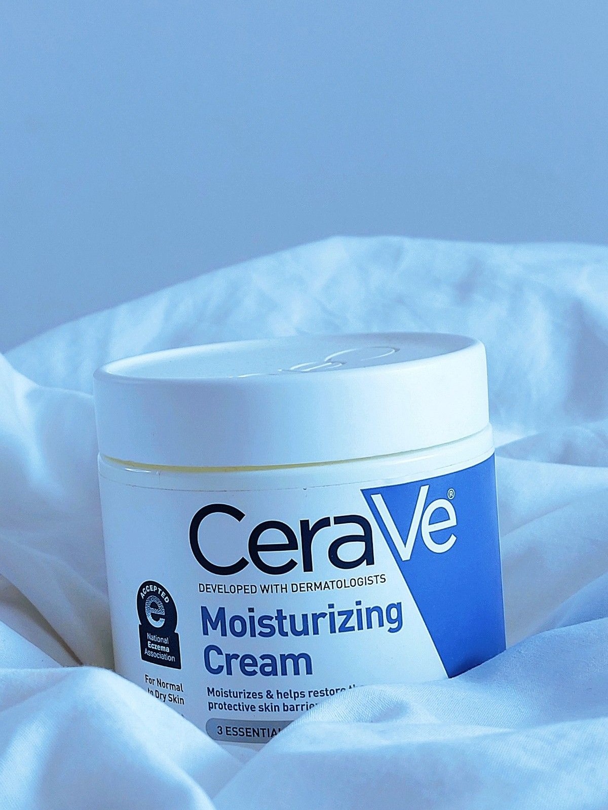 CeraVe Moisturizing Cream jar nestled in white bedding lit in blue tones