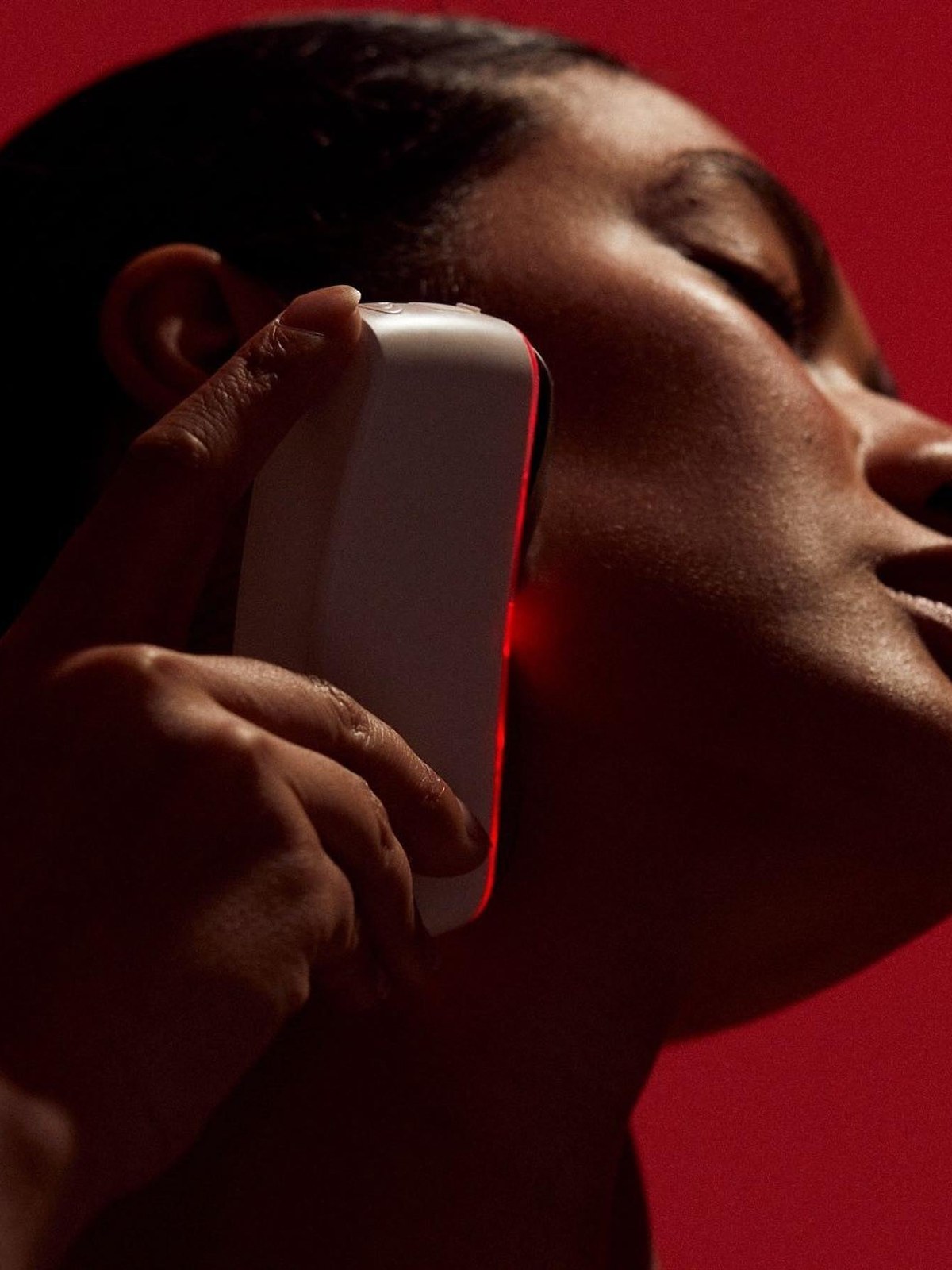 Model using a red-lit facial device in a dramatic close-up against a red backdrop