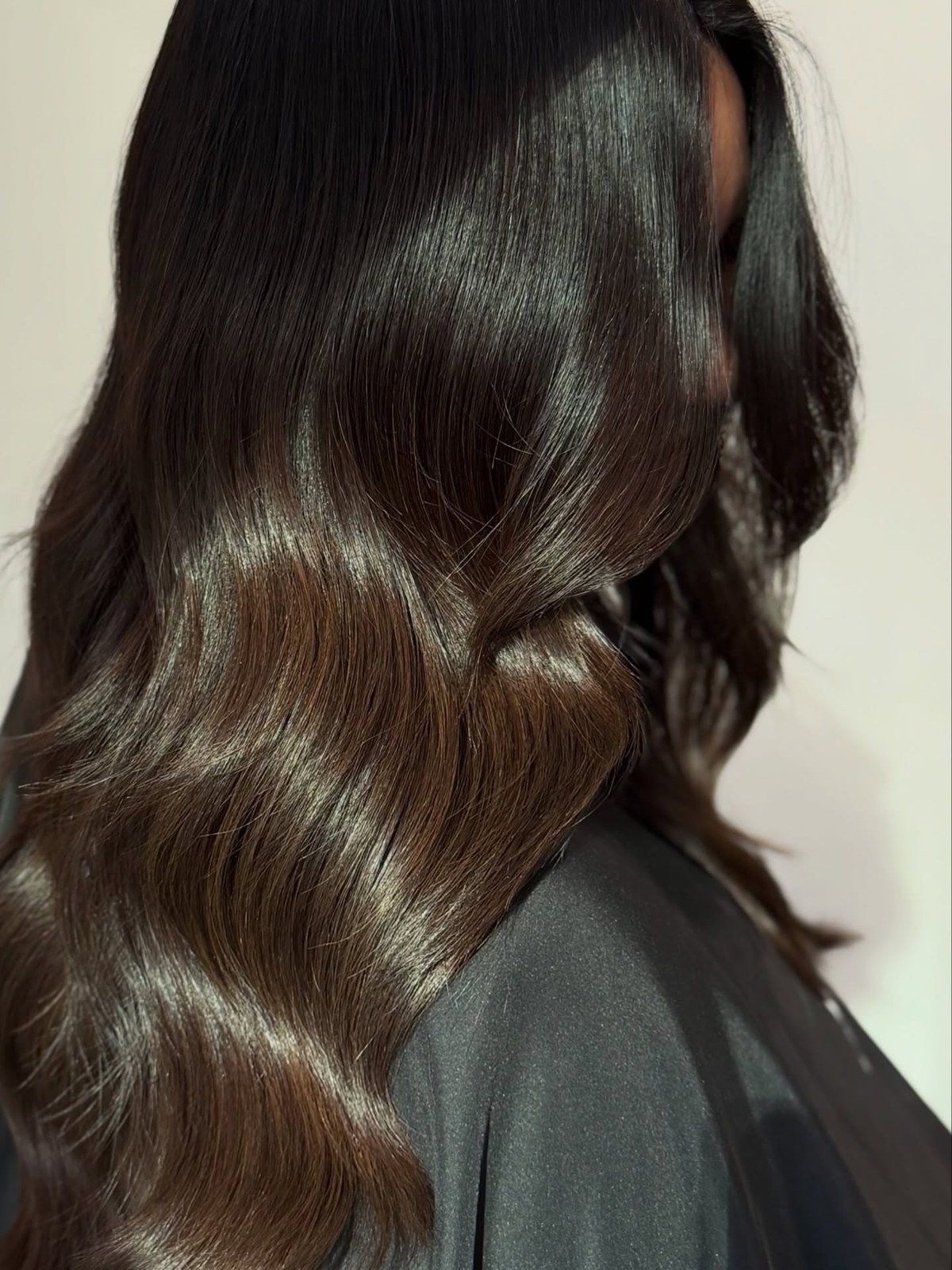 Side view of long, glossy, dark brown waves with warm chestnut ends, worn over a black salon cape