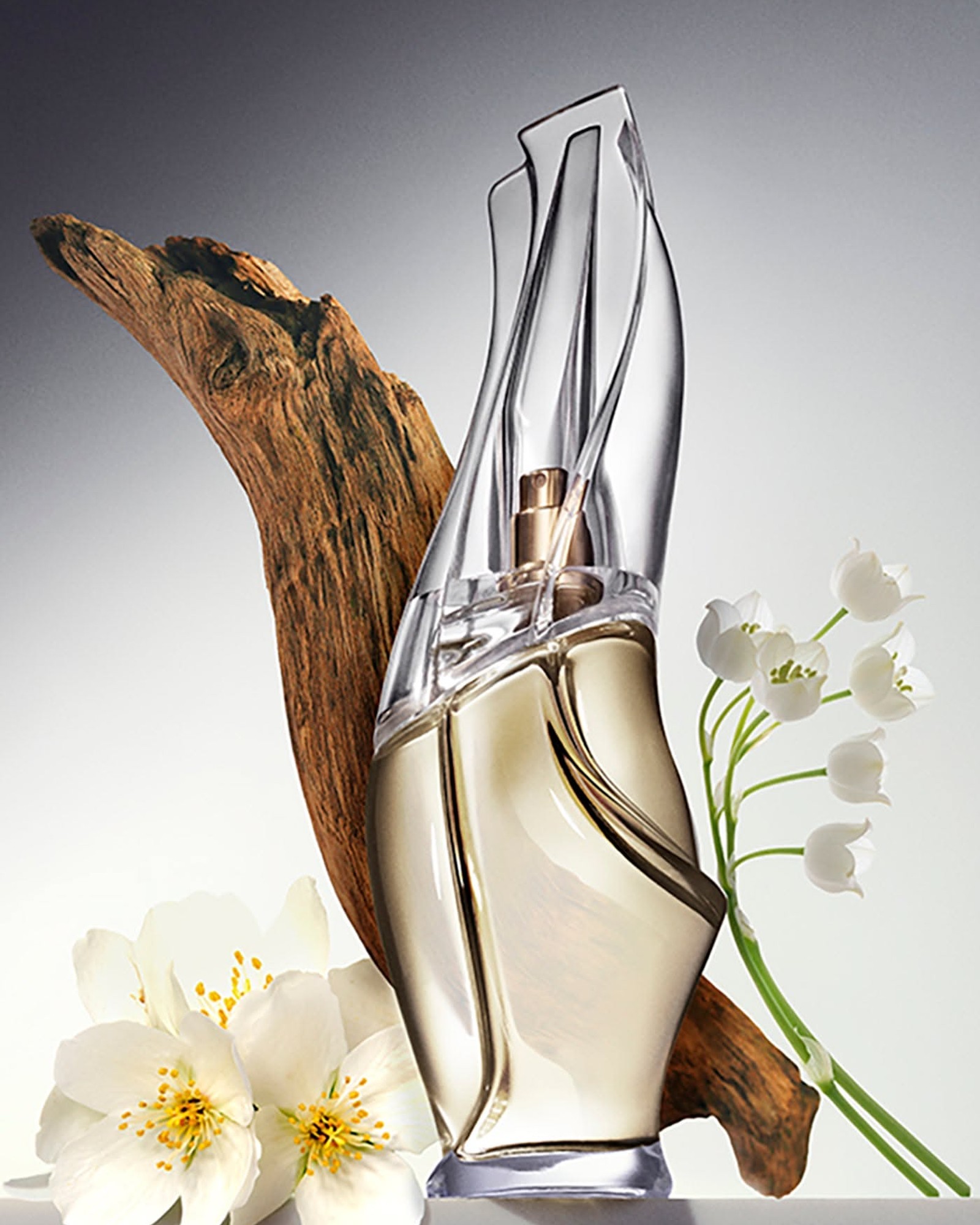 Cashmere Mist perfume bottle leaning against a piece of driftwood, with white jasmine blossoms and lily of the valley beside it