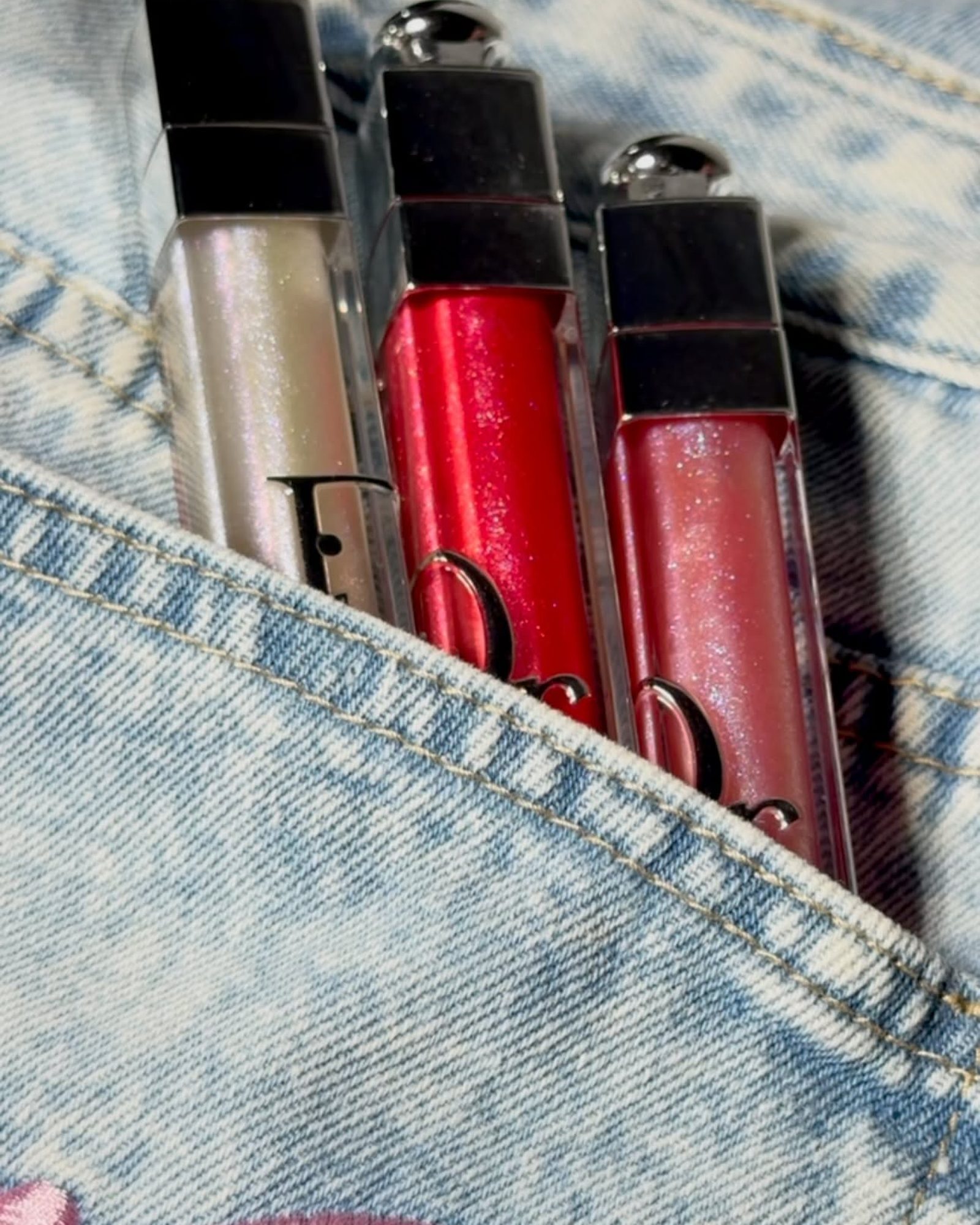 Three Dior Addict Lip Maximizer glosses in pearl, red and rose shimmer tucked into the pocket of a light denim jacket