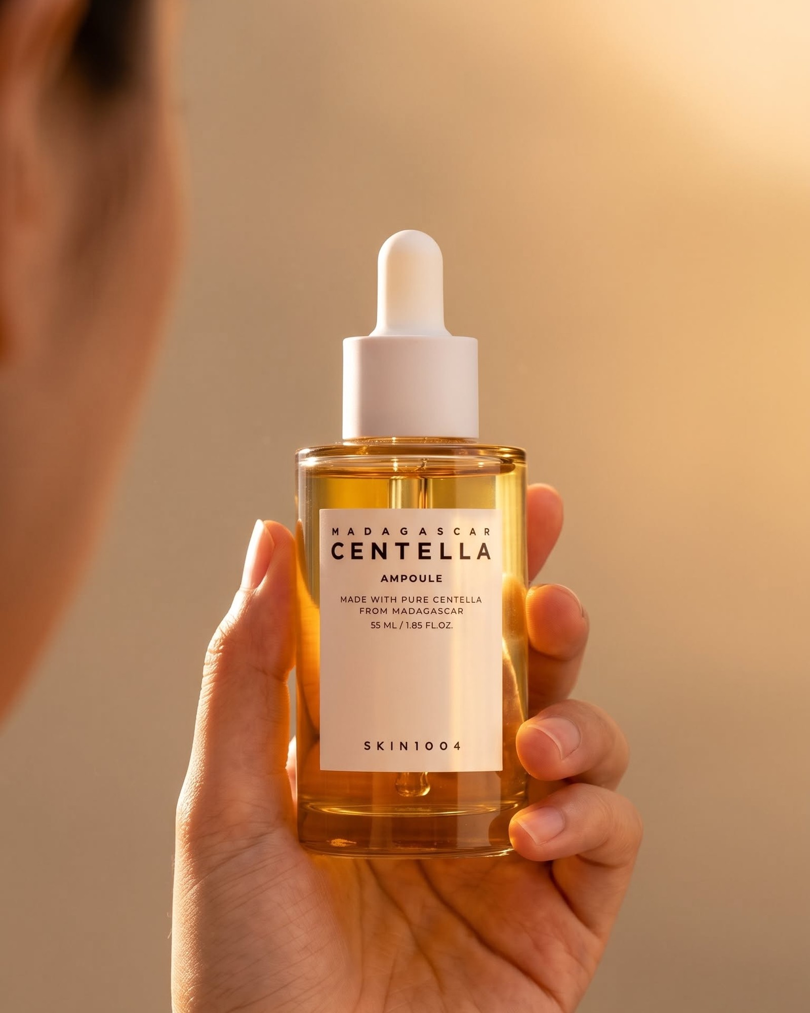 Hand holding a SKIN1004 Madagascar Centella Ampoule 55 ml bottle in warm golden light with a blurred face at the edge