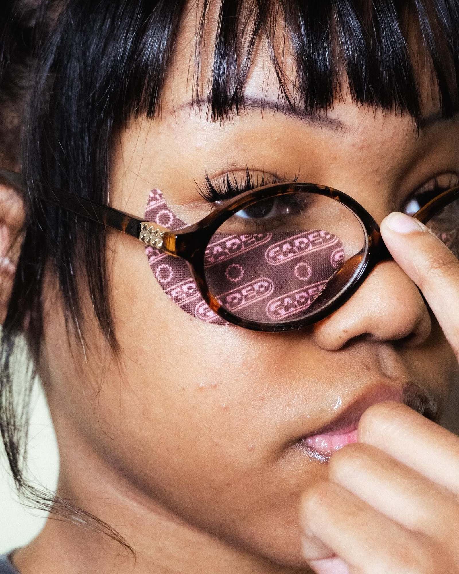 Close-up of a woman with black bangs and tortoiseshell glasses wearing a dusty pink Faded patch under one eye, fingers touching her nose.