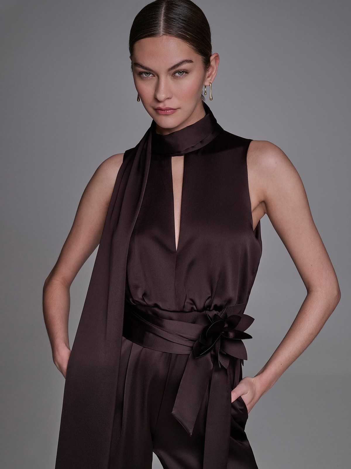 Model in a chocolate satin jumpsuit with a scarf neck and waist bow