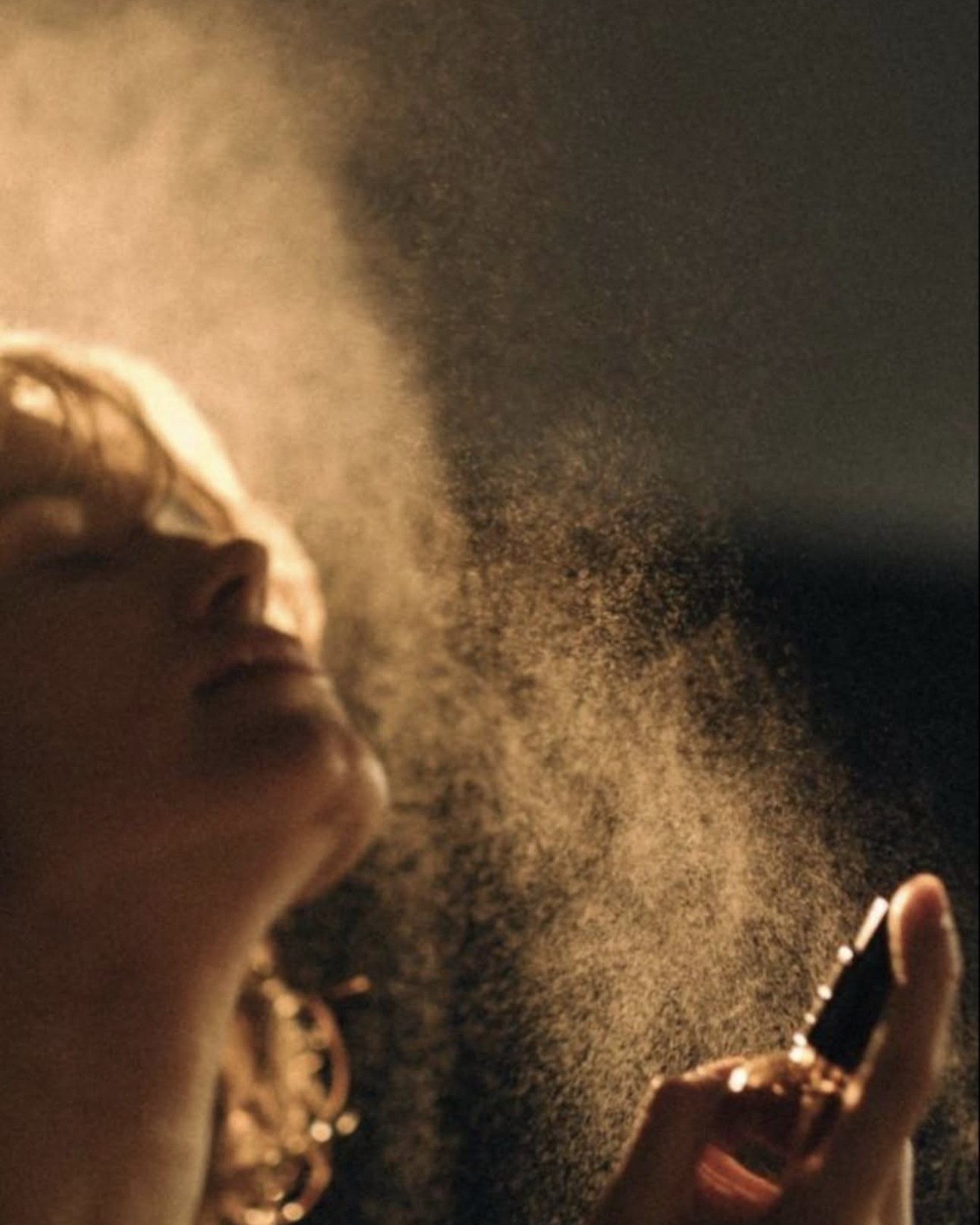 Woman tilting her head back in warm low light as a golden perfume mist sprays from a small bottle in her hand