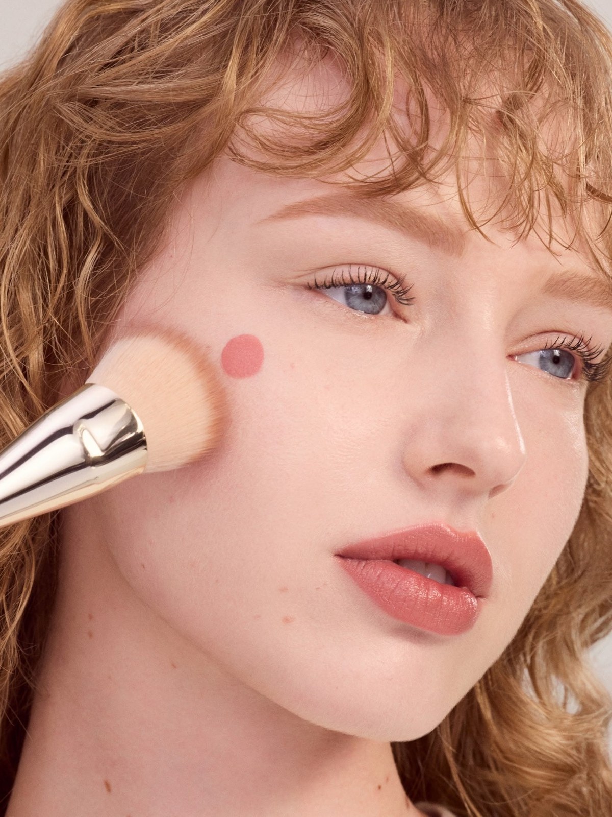 Curly-haired model applying pink blush with a brush