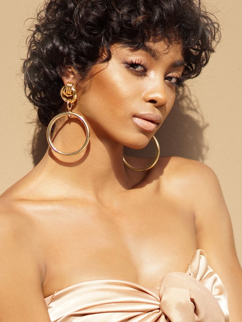 Model in a satin top wearing oversized gold hoop earrings against a beige studio background.