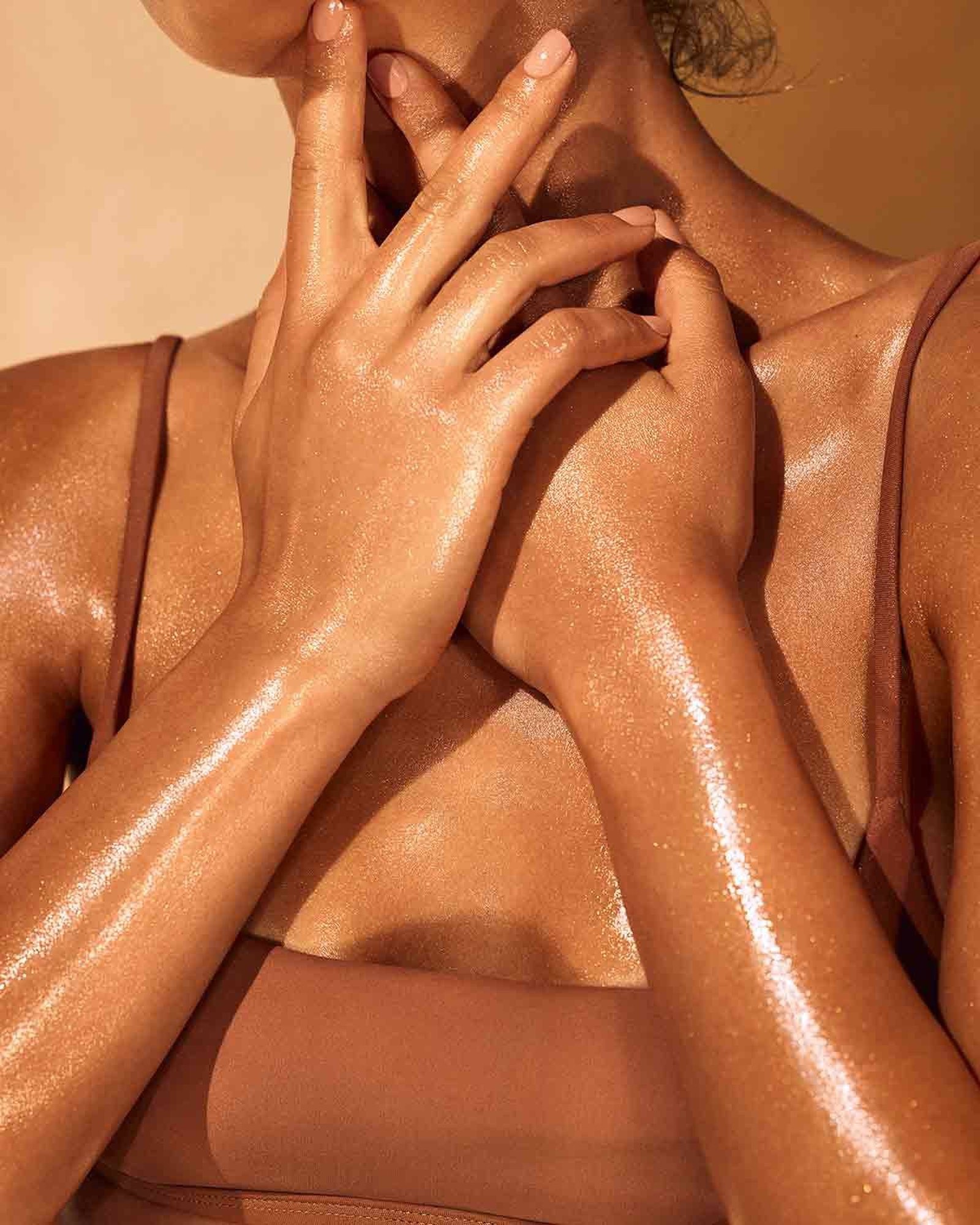 Close crop of glistening tan skin on hands, neck and arms with a light shimmer, wearing a brown bralette
