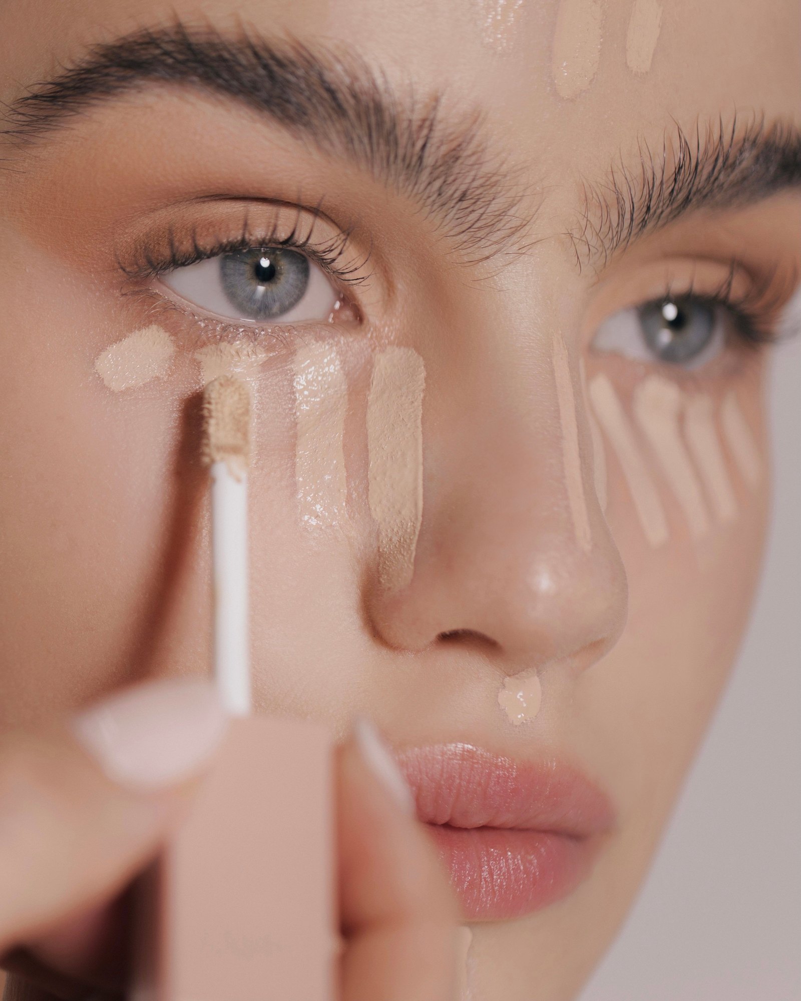 Extreme close-up of a model's eye area with several stripes of concealer in different shades and a white applicator held in front