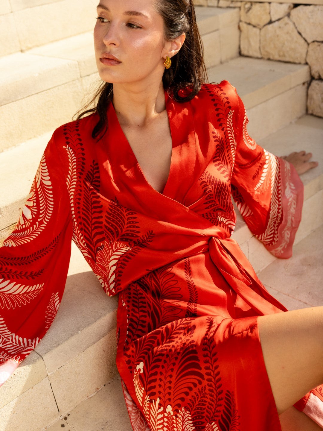 Model in a red patterned wrap dress seated on pale stone steps