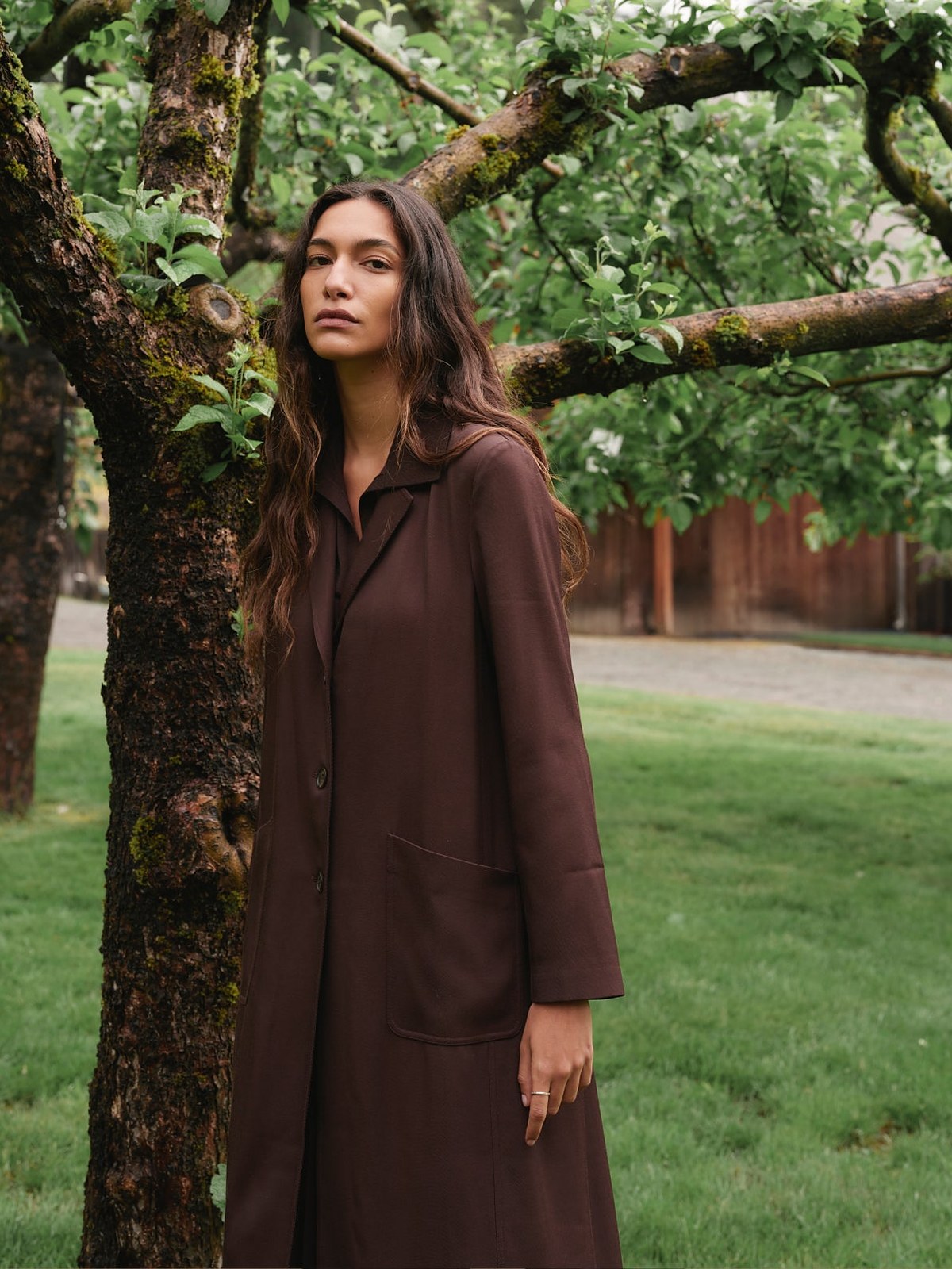 Model in a brown wool long coat standing under an apple tree