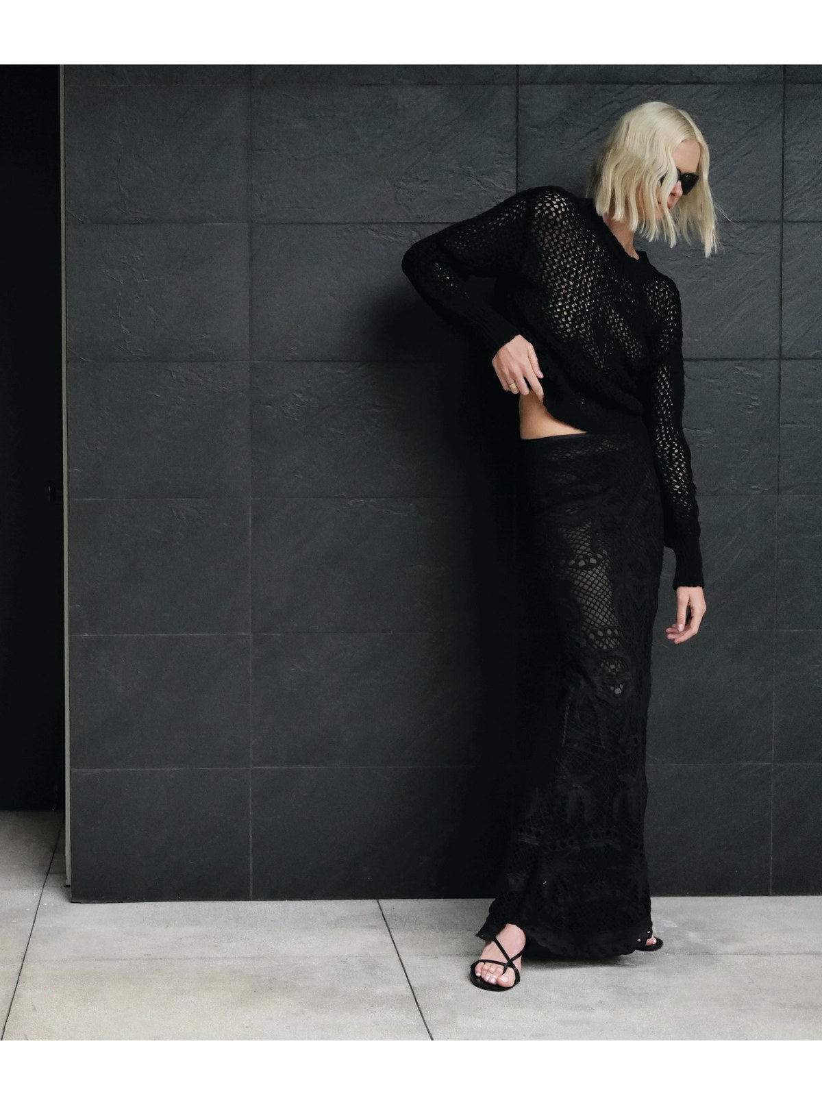 Blonde model in a black open-knit look against a dark tile wall
