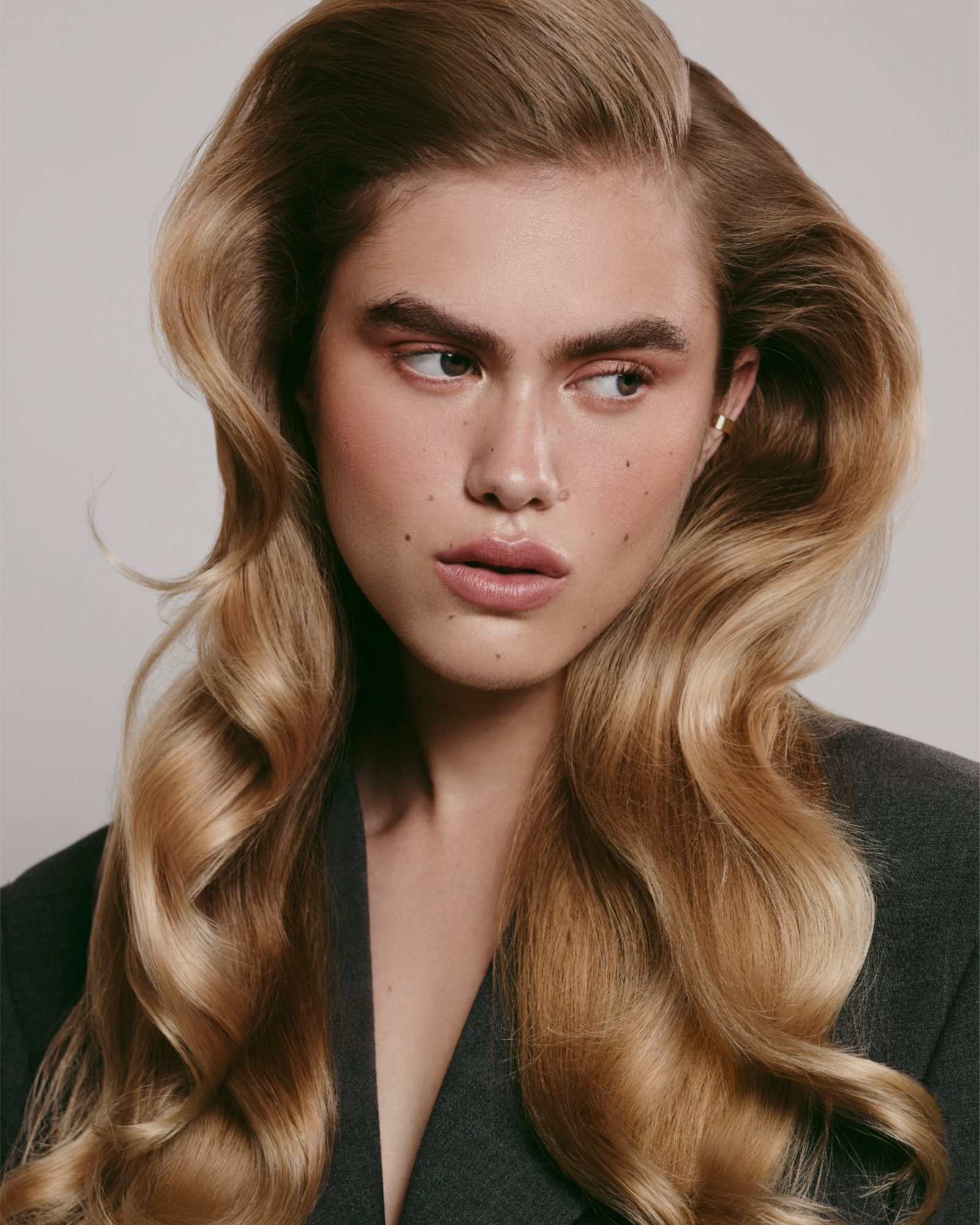 Woman with long, glossy caramel blowout waves in a dark blazer against a grey backdrop