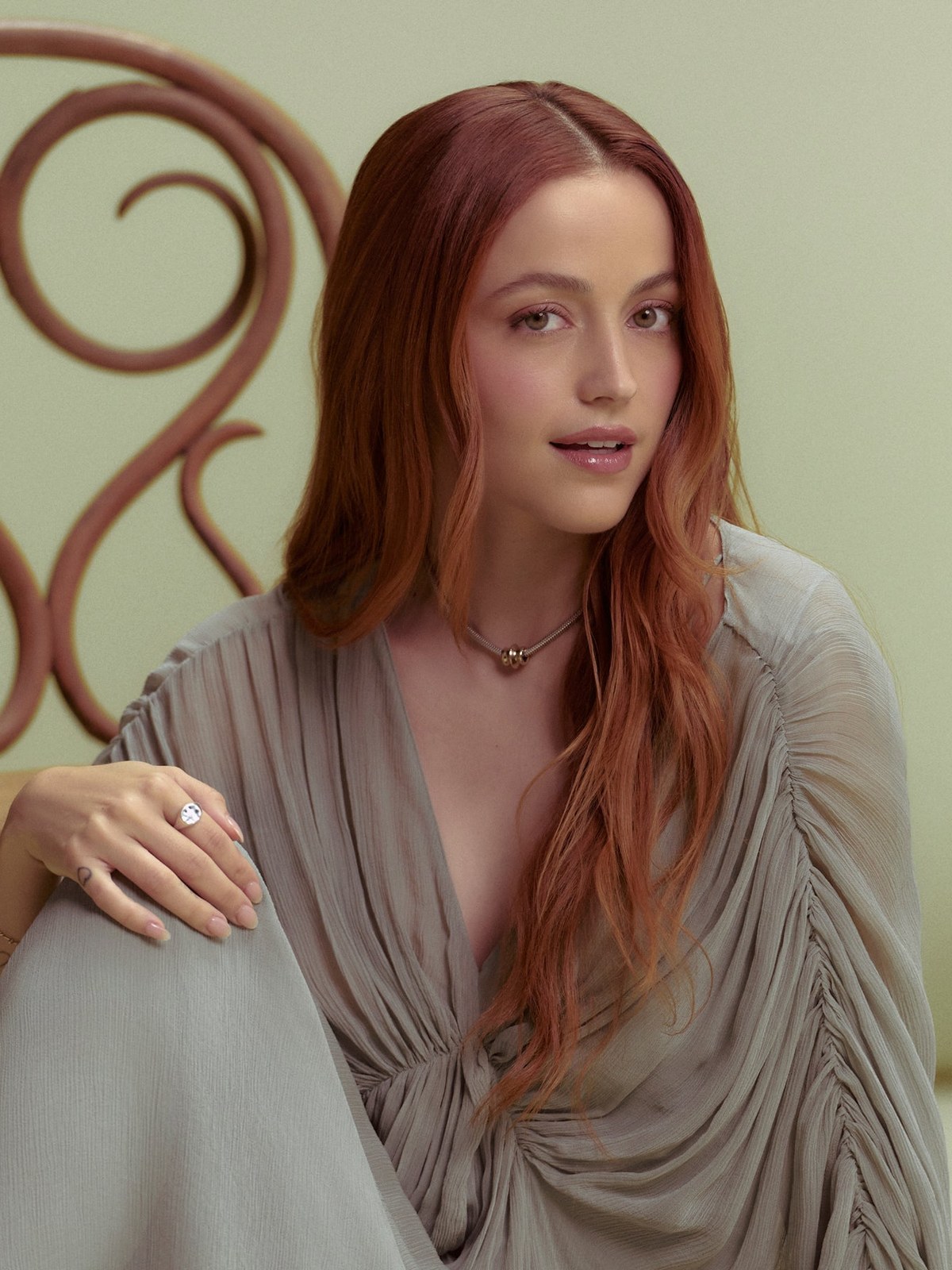 Red-haired creator seated in a pale draped dress in a soft green setting