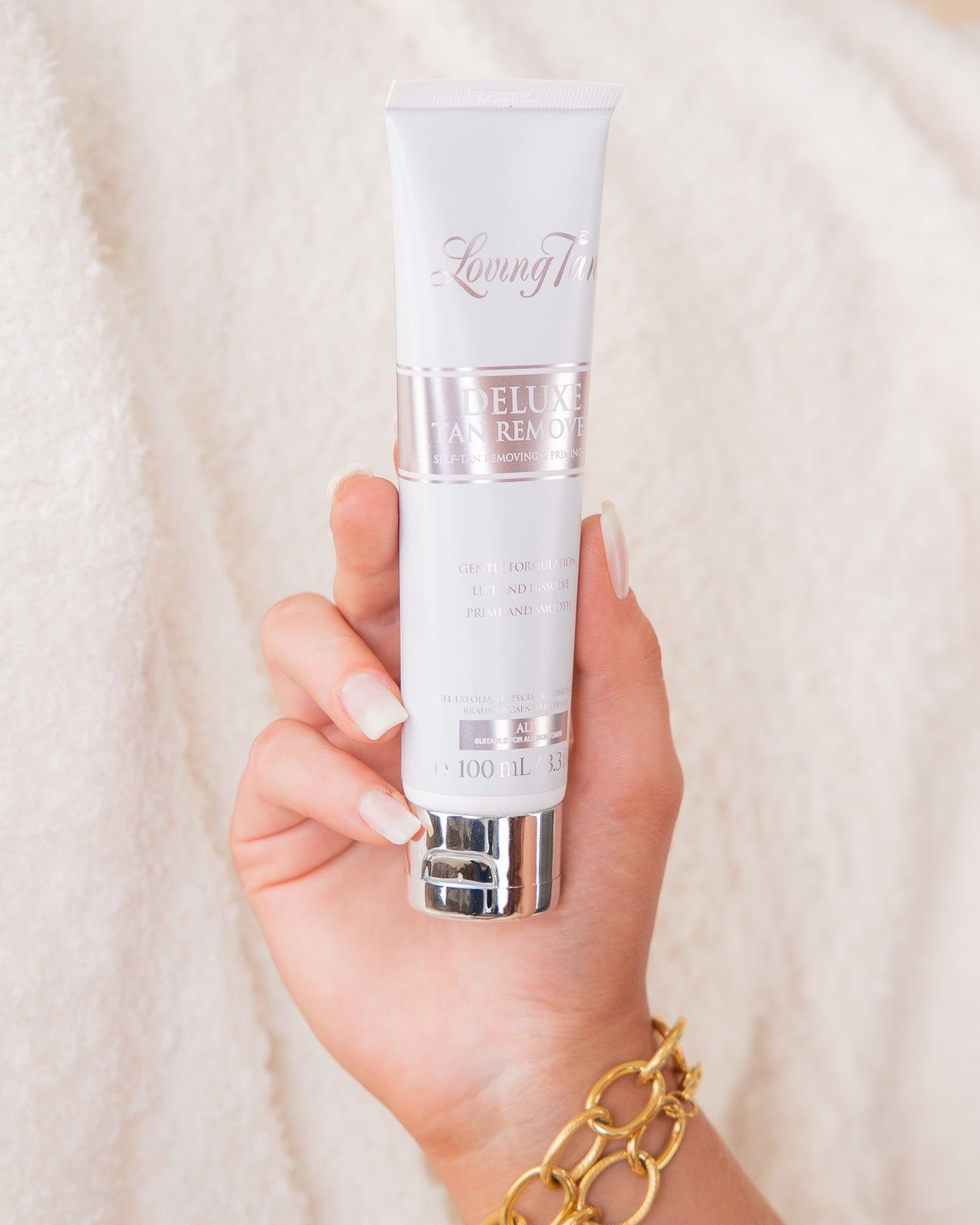 Hand with pale manicured nails holding a white and silver tube of Loving Tan Deluxe Tan Remover against a cream fluffy blanket, with a gold chain bracelet