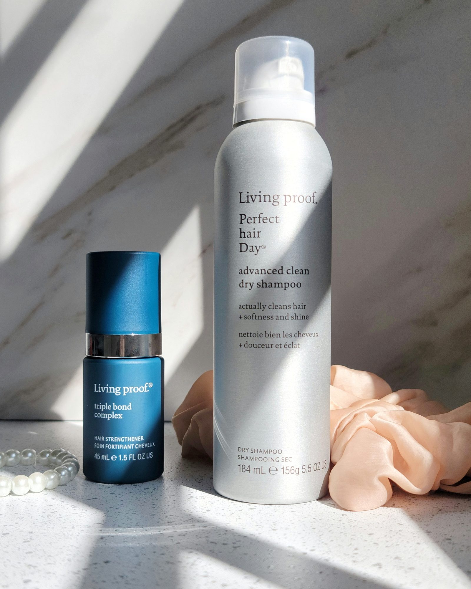 Living Proof triple bond complex bottle beside a Perfect hair Day advanced clean dry shampoo can on a marble counter with a peach scrunchie